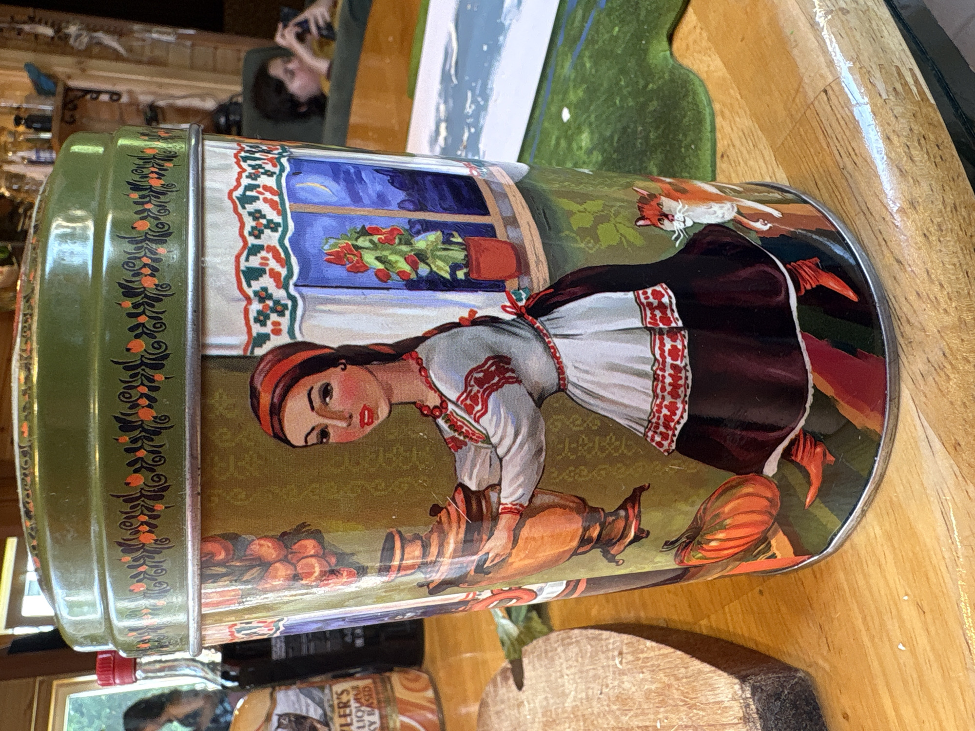 The image shows a close-up of a cylindrical tin decorated with a folk art scene. A woman in traditional dress, holding a samovar, stands in front of a window with a potted plant and a night sky visible. A fox is depicted near the bottom of the tin. The tin sits on a wooden surface, likely a table, with a wooden cutting board to the left. In the background, a person is visible looking at a phone in a dimly lit room. The scene is likely indoors, possibly in a home, and there is no specific indication of the location being Lyady, Russia.