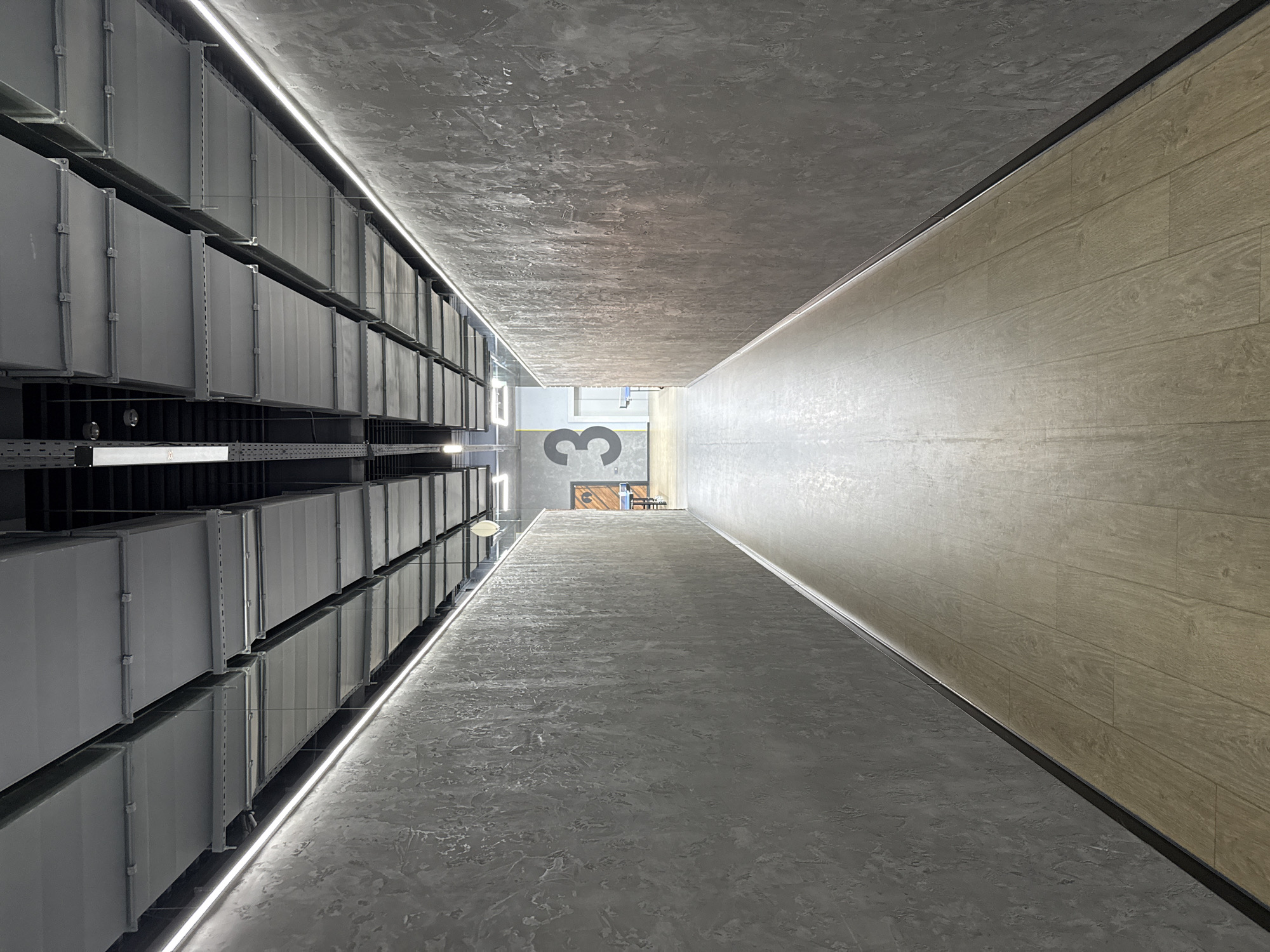 The image is an interior shot of what appears to be a corridor or hallway, taken from a unique perspective. The location is unknown, but the user specified Votkinsk, Russia.
The left side of the image features a series of gray, metallic structures, possibly ductwork or storage units. These are arranged in a linear fashion, receding into the distance. Underneath them, there are black metal beams.
The ceiling is a textured, gray material. The right wall has a wood grain pattern.
In the distance, at the end of the hallway, there is a large number "3" painted on the floor. There is also a door or opening leading to another space.
There are LED lights or light strips running along the edges of the ceiling and walls, providing illumination. The overall lighting is bright and even.
There are no people visible in the image. The scene appears to be a modern or industrial setting.