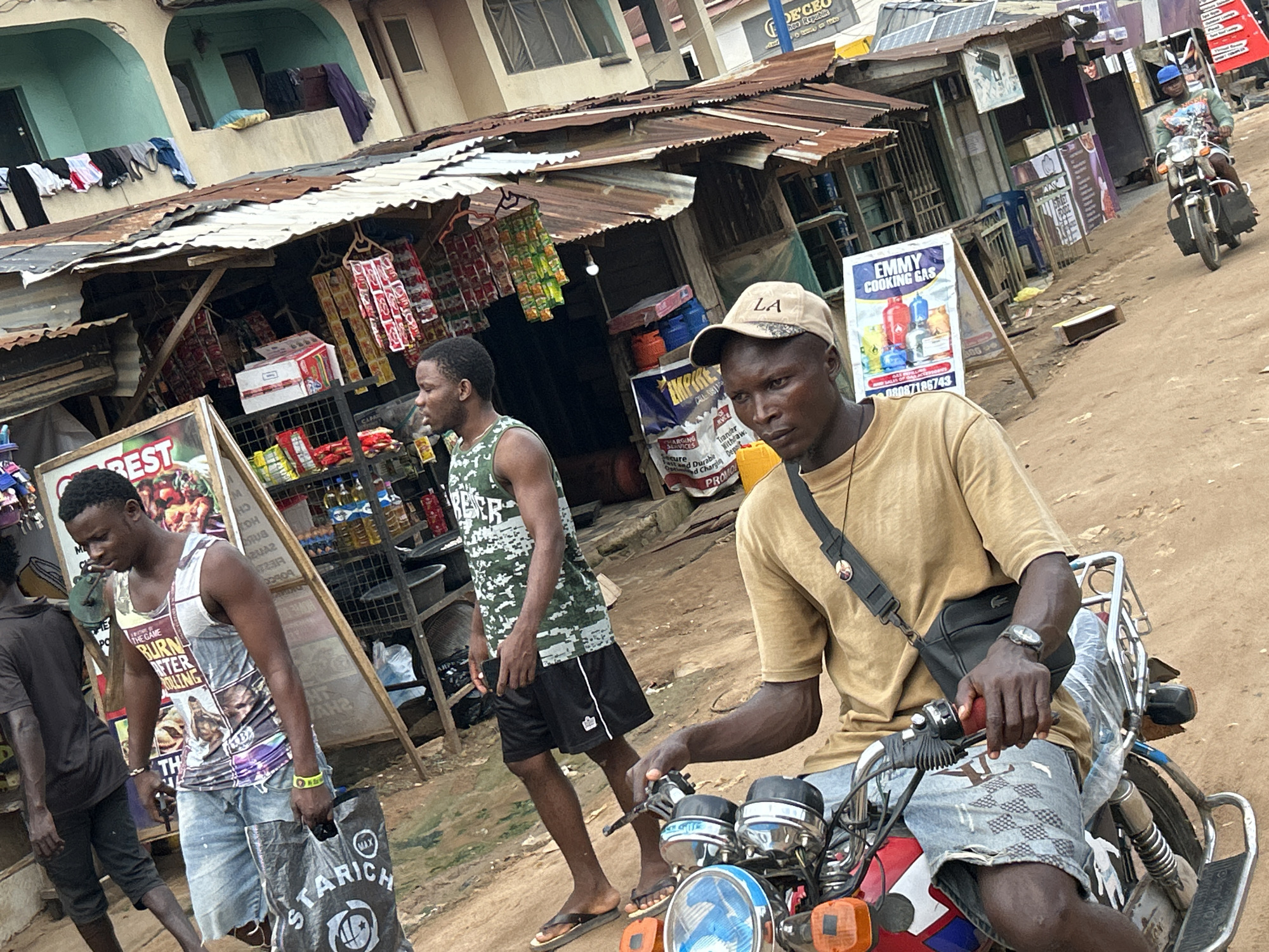 In Ihiagwa, Nigeria, a bustling street scene unfolds in front of a row of market stalls. The foreground is dominated by two men, one on a red motorcycle and the other standing beside him. The man on the motorcycle, wearing a tan t-shirt, denim shorts, and a beige baseball cap, has a black messenger bag slung across his shoulder and is holding the handlebars. Beside him, a man in a camouflage tank top and black shorts walks with a large black shopping bag in his hand. Behind them, a motorcycle carrying a rider in a green and white shirt passes by.
The market stalls themselves are constructed with corrugated iron roofing and wooden frames, displaying various goods including bottled drinks, packaged foods, and clothing. A sign advertising "EMMY COOKING GAS" is prominently displayed. Laundry hangs from a line above one of the stalls. The ground is covered in dusty, reddish-brown earth, indicative of the local environment. The overall atmosphere is one of everyday commerce and activity in a Nigerian town.
