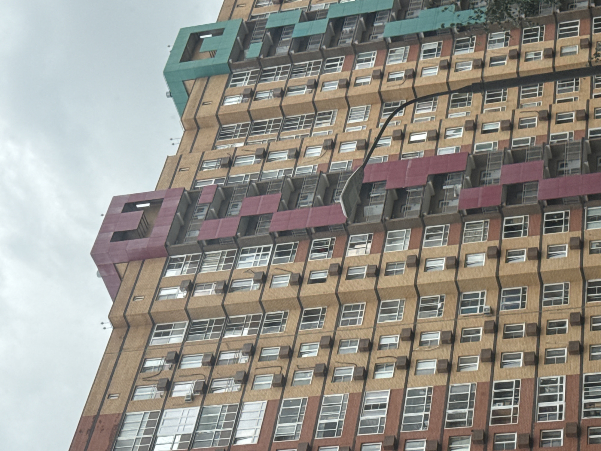 This is a medium-shot, eye-level, outdoor, daytime photograph of a tall, modern apartment building in Misaylovo, Russia. The building is constructed with tan-colored brick and features a prominent vertical band of dark pink and another of teal running up the facade. Numerous windows and balconies are visible, along with multiple air conditioning units mounted on the exterior. A black lamppost curves into the frame from the right side. The sky above is overcast and gray, suggesting a cloudy day. No people are visible in the image.