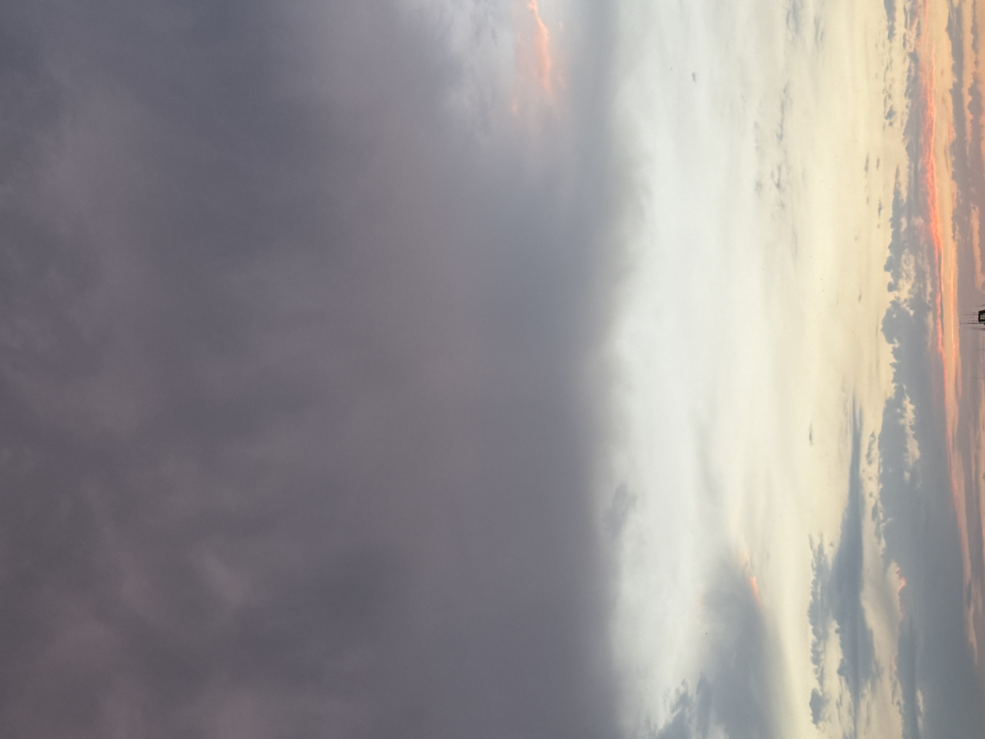 The image shows a wide shot of a sky filled with clouds, with a hint of sunset colors visible at the bottom. The clouds are a mix of dark grays and lighter whites, with streaks of orange and pink near the horizon. There are no people, subjects, or objects clearly identifiable in the frame, except for what appears to be the top of a structure at the very bottom edge of the image. The scene depicts an atmospheric sky, likely taken during the late afternoon or early evening. The location is stated to be Bishkek City, Kyrgyzstan, but the image itself does not contain any specific geographical markers that would confirm this.