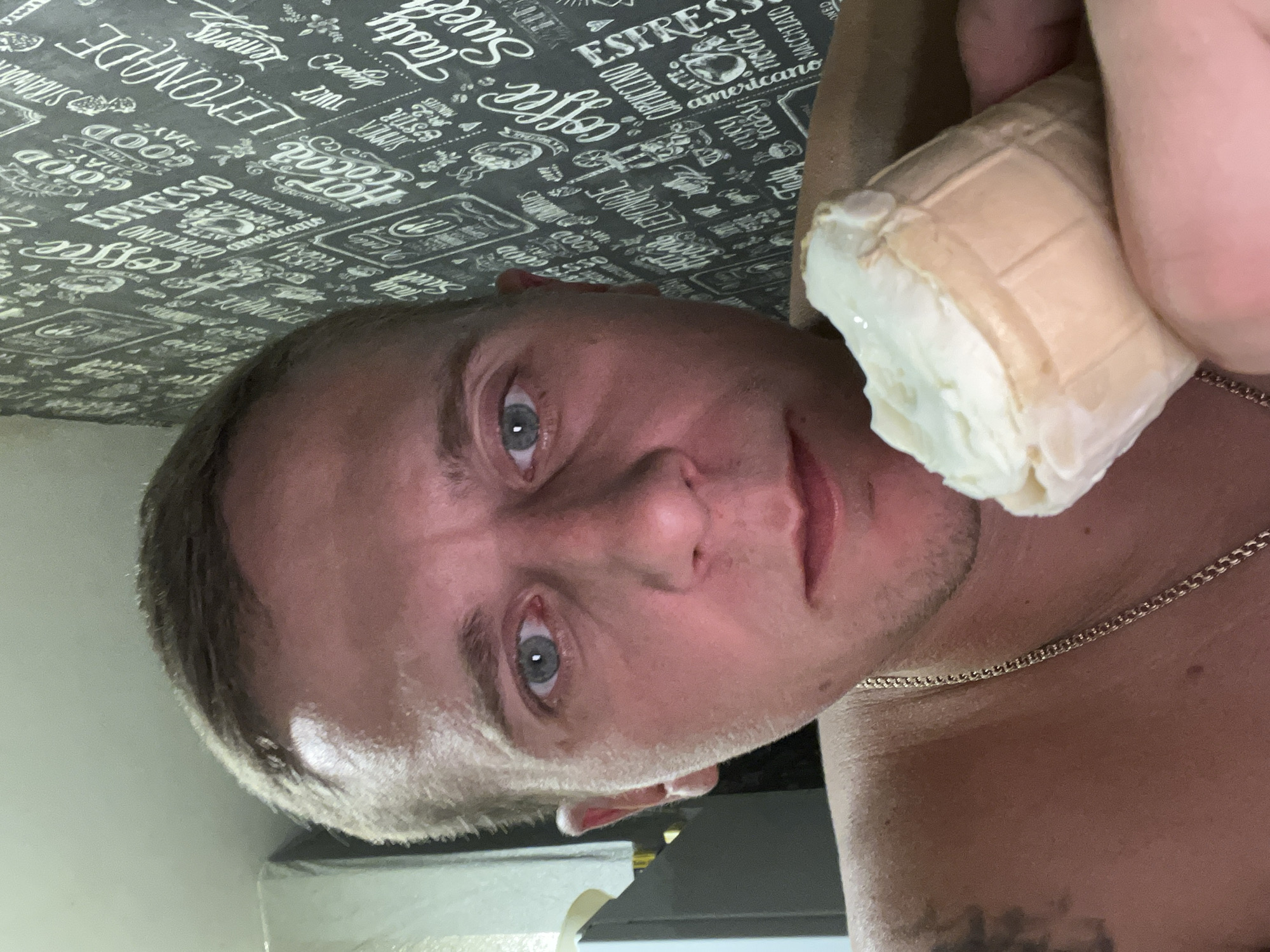 A selfie taken indoors, showing a man holding an ice cream cone to his mouth. He has fair skin, short brown hair, and blue eyes. He is wearing a gold chain. The background is a wall covered with black and white wallpaper that features text and graphics related to coffee and drinks. The setting appears to be domestic, possibly an apartment in Minsk, Belarus, based on the context provided.