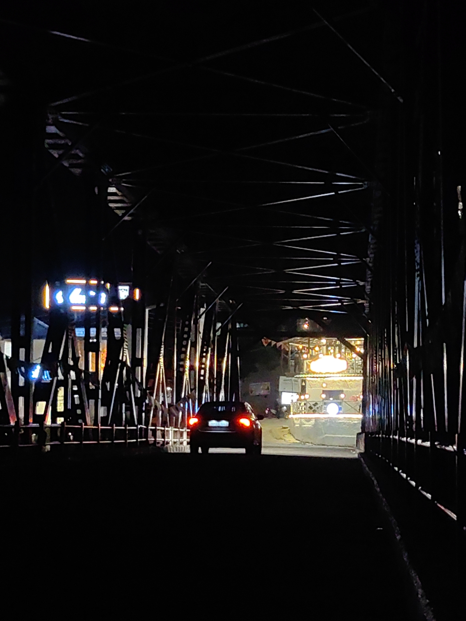 The image depicts a night view from within a bridge structure, possibly in Kulang, India. A dark-colored car is visible driving away from the viewer on the road beneath the bridge. The car's brake lights are illuminated red. The bridge's metal framework is prominent, with various beams and supports creating a dark, skeletal appearance. On the left side of the frame, illuminated signs with blue and orange lights are present. Further ahead, beyond the immediate bridge structure, a brightly lit area with what appears to be a stage or event setup is visible, featuring numerous lights and some small structures. The overall scene is characterized by the contrast between the dark, structural elements of the bridge and the illuminated areas ahead and on the left. The road surface is visible beneath the car.