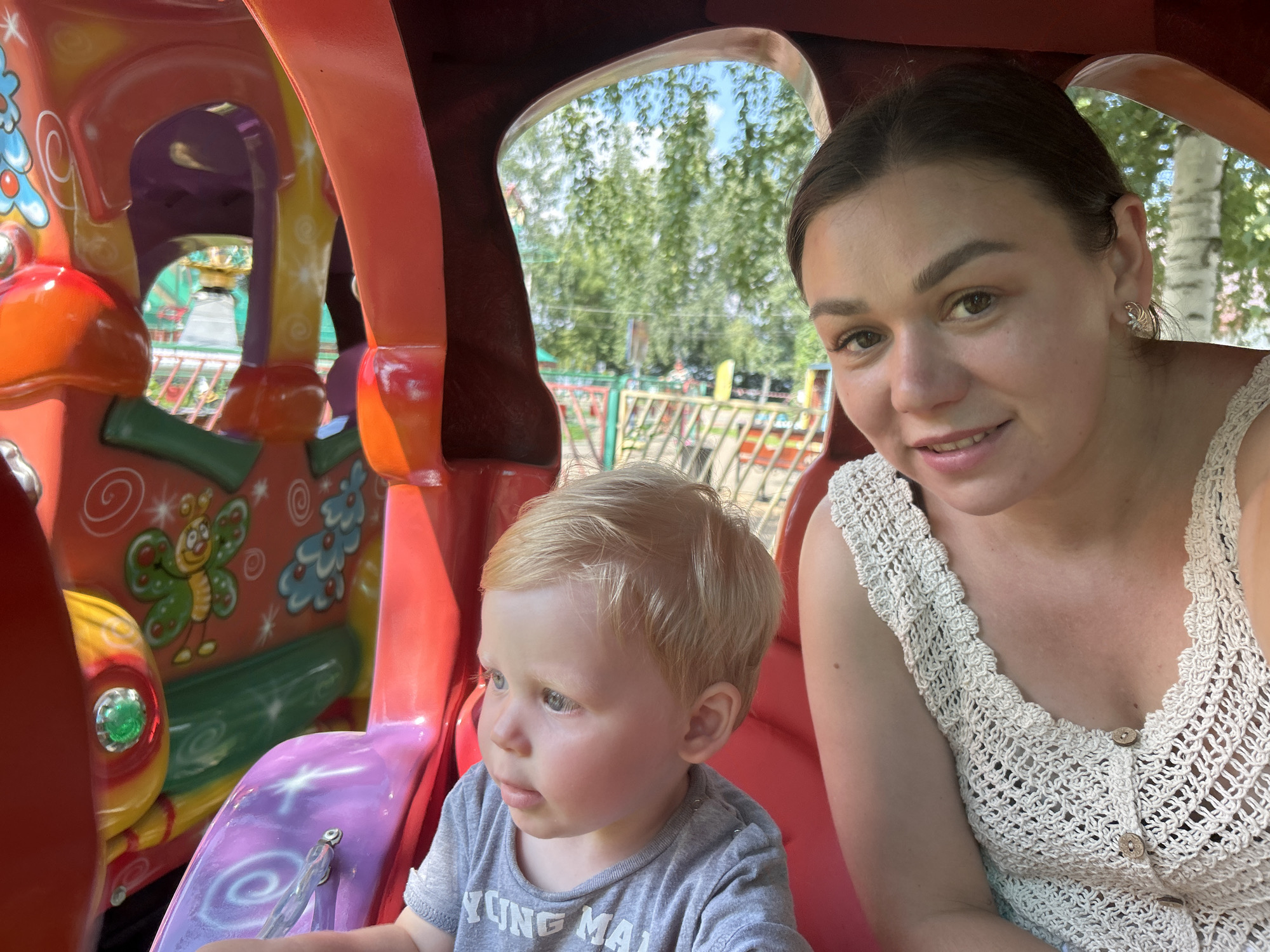 A young woman and a toddler are seated inside a colorful amusement park ride. The ride appears to be a vehicle with a red exterior and purple and orange interior. The woman, with dark hair pulled back and a slight smile, wears a cream-colored crocheted top. The toddler, with blond hair and blue eyes, is looking off to the side. The background visible through an opening in the ride shows trees and a fence, suggesting an outdoor park setting. The image is a close-up, with the ride's interior decorations, including a butterfly motif, visible on the left. The setting is likely an amusement park in the city of Chaikovsky, Russia, given the provided context.