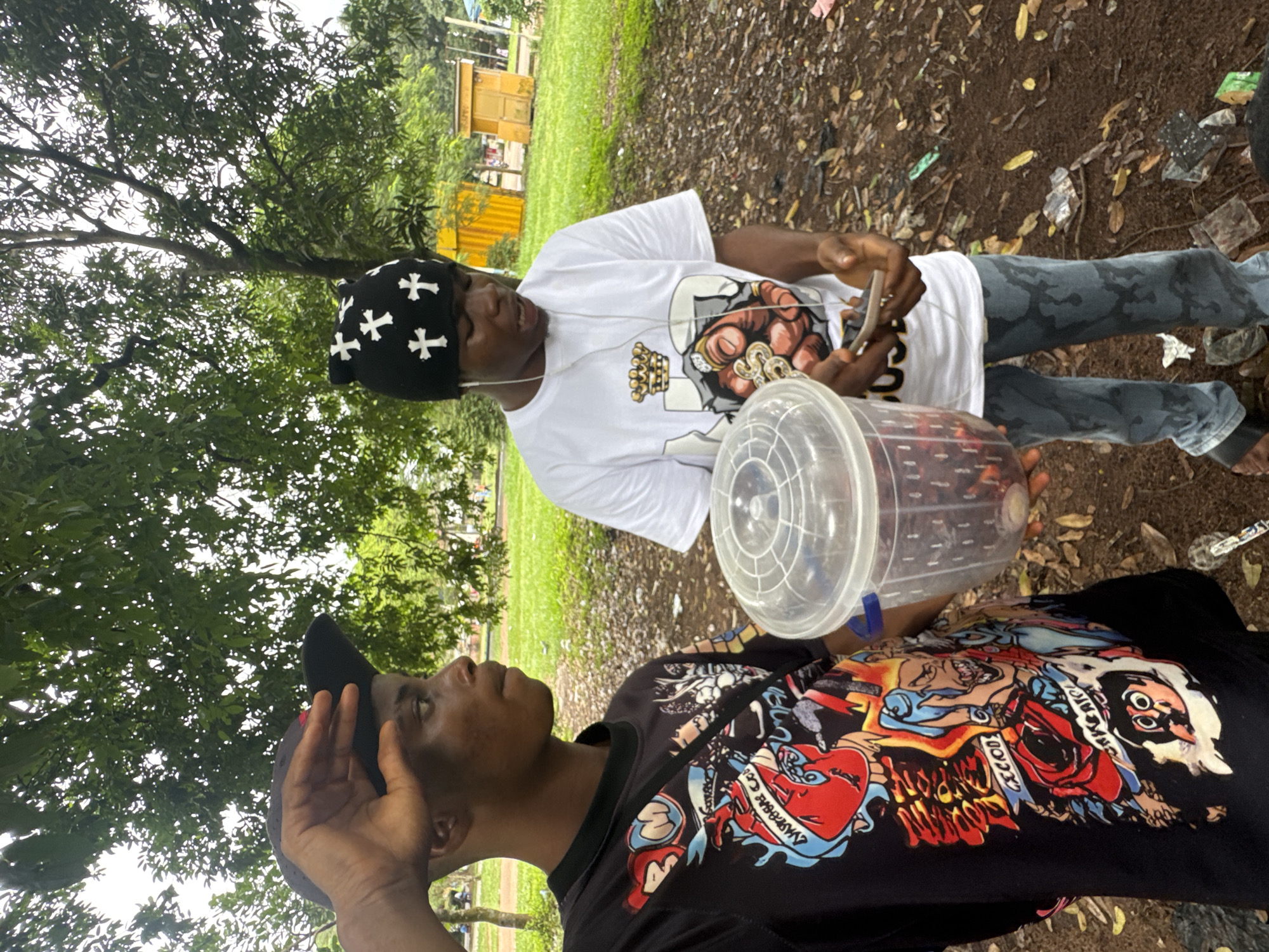Two young men are standing outdoors in what appears to be a park or open field in Ihiagwa, Nigeria. The man on the left is wearing a black t-shirt with a colorful, graphic print and a black baseball cap. He is raising his hand to his forehead, as if shielding his eyes from the sun. He is holding a clear plastic container filled with dark red, possibly fruit, in his left hand. The man on the right is wearing a white t-shirt with a graphic design and a black beanie with white crosses. He is looking down at his phone, which he is holding in his hands, and appears to be speaking. He has earbuds in his ears. The ground is covered with dirt, leaves, and some scattered litter. Trees are visible in the background, along with some yellow structures that could be temporary shelters or storage units. The overall lighting suggests an overcast day.