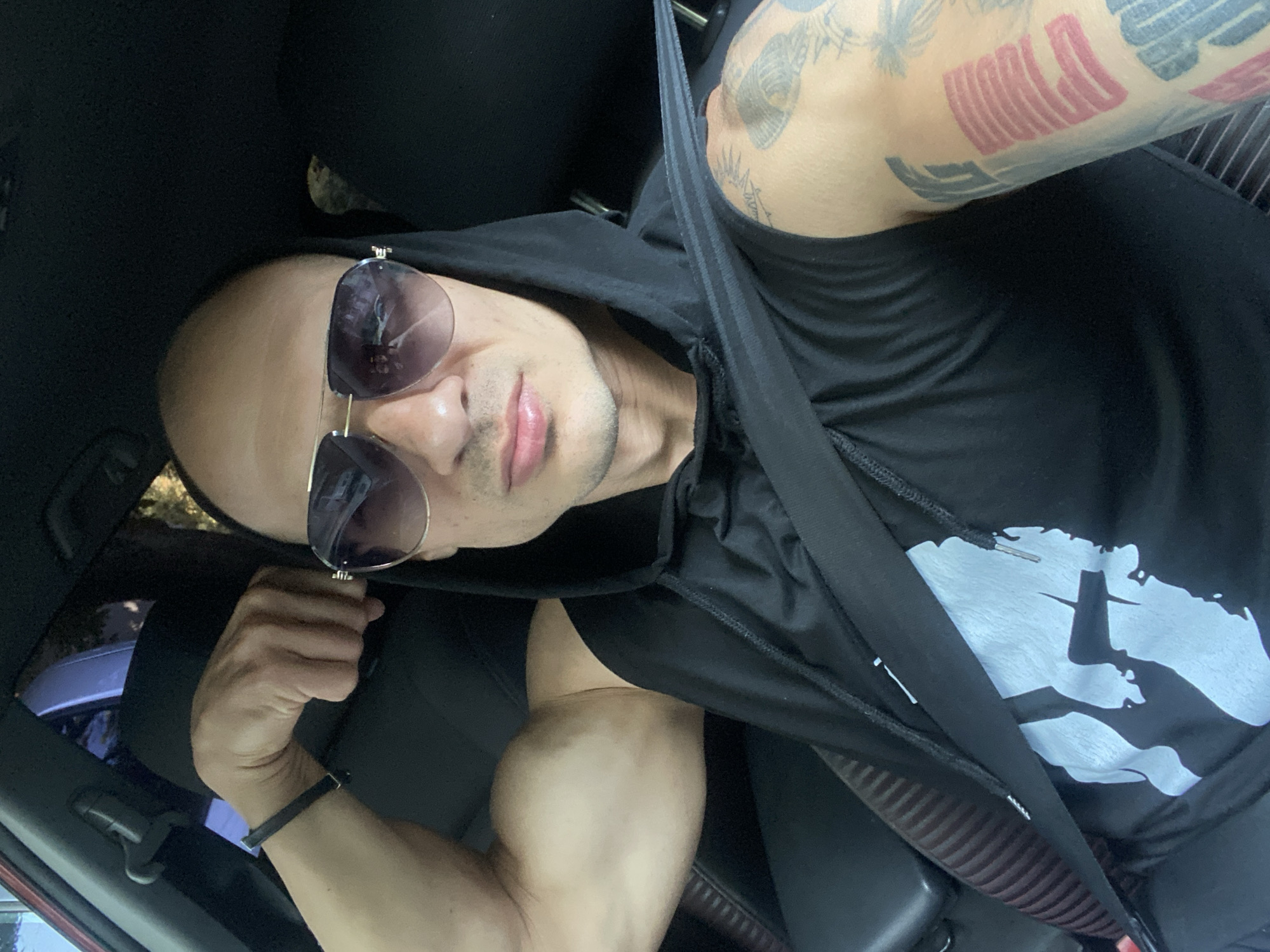A man is sitting in the driver's seat of a car. He is wearing a black hooded shirt with a white graphic on the front and sunglasses. His left arm is raised and flexed, showing his bicep. A seatbelt is fastened across his chest. There are visible tattoos on his right arm. The interior of the car appears to be black. The lighting suggests it is daytime. The background outside the car is blurred and shows trees.