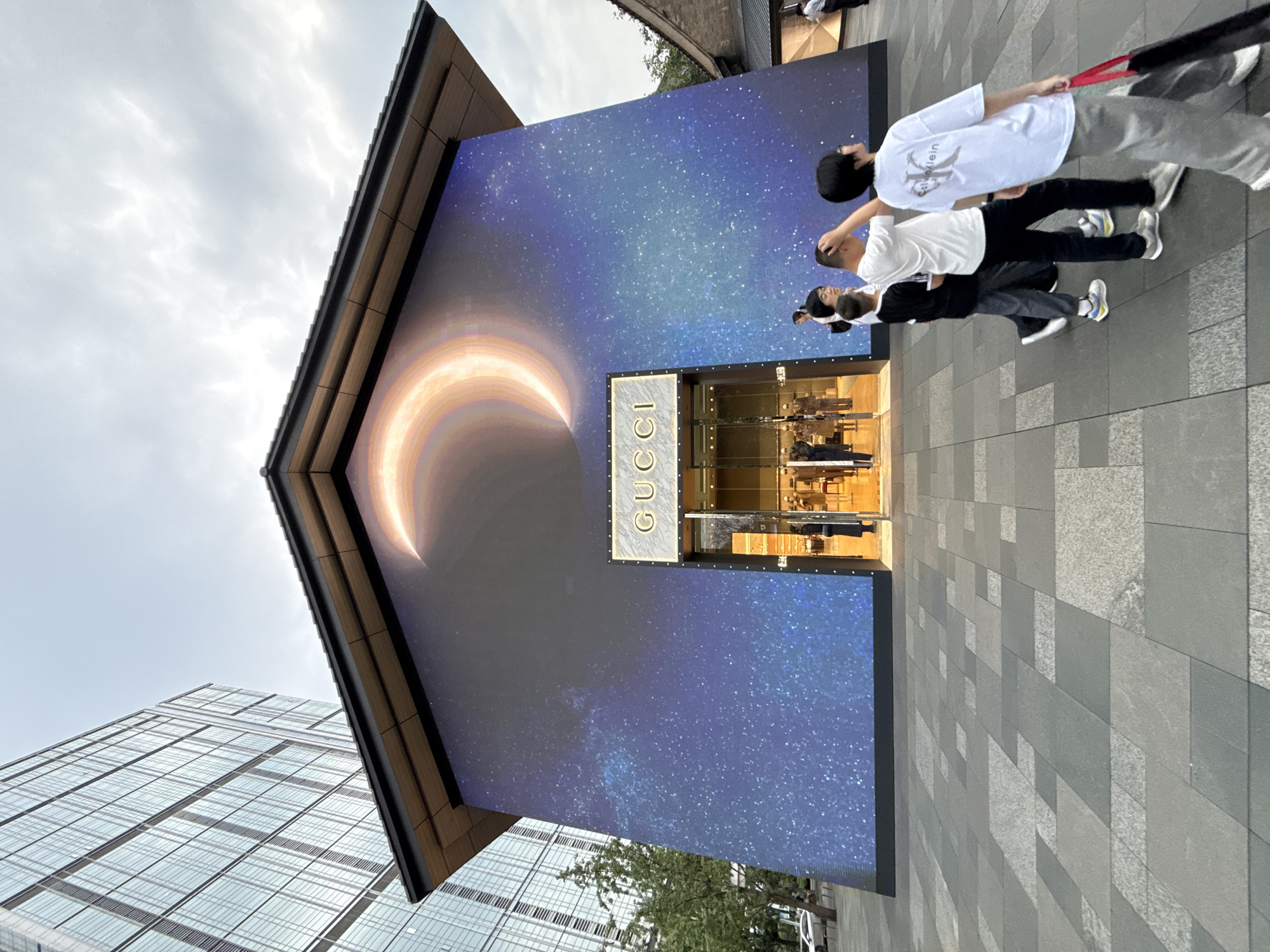 An outdoor, eye-level shot captures a Gucci store in Jinjiang District, China, featuring a large LED facade displaying a bright, glowing crescent moon against a vibrant blue and purple starry night sky. The building has a traditional dark tiled roof with subtle upward-curving eaves, contrasting with the modern LED screen. A brightly lit glass entrance in the center of the facade displays the "GUCCI" logo above it, with mannequins visible inside.
In the foreground, a paved pedestrian plaza, composed of varying shades of grey square and rectangular tiles, extends from the viewer towards the store. Several individuals are walking on this plaza from left to right. On the far right, a person wearing a white t-shirt with "Calvin Klein" printed on the back and grey pants holds a black bag with a red strap. To their left, another person in a white t-shirt and dark pants raises their right hand to their head. Further left, a third person in a black shirt and grey pants walks alongside. Other individuals are partially visible or less distinct in the foreground and inside the store.
To the left of the Gucci store, a tall, modern glass skyscraper with numerous windows is visible, reflecting the sky. A small tree with green foliage stands between the store and the skyscraper. To the right of the Gucci store, a portion of another building with a dark roof and lighter walls, appearing to be of traditional architectural style, is visible. The sky above is overcast with light, diffused clouds.