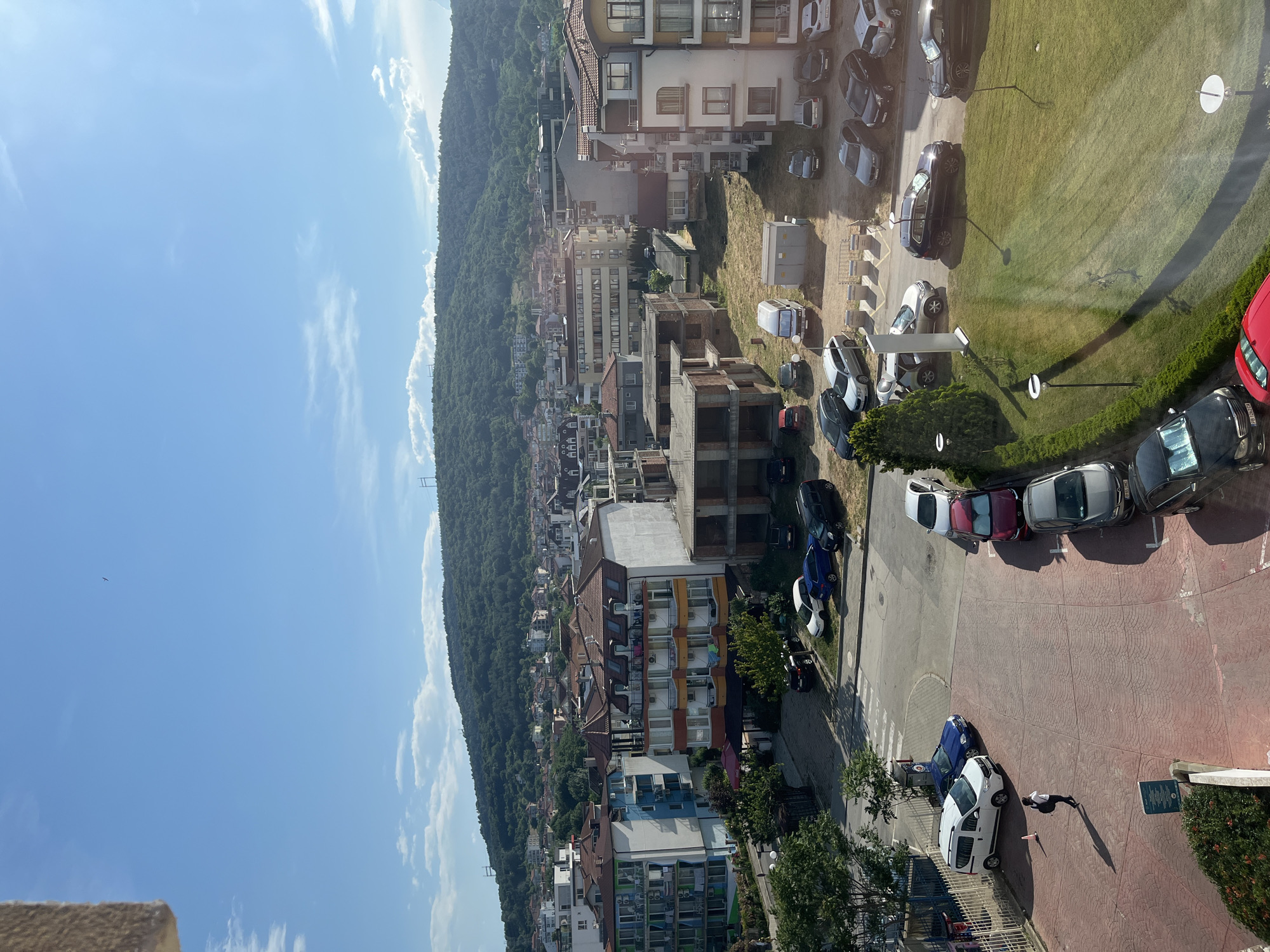 An aerial view captures a bustling scene in Obzor, Bulgaria, under a clear blue sky dotted with wispy clouds. In the foreground, a woman with dark hair, wearing a white top and dark pants, walks across a red brick paved area, casting a long shadow. She carries a dark backpack. A white van is parked to her left.
Further into the scene, cars are parked in various lots. A white van is positioned near a grassy area, adjacent to a small structure. Several cars are lined up along a road and in parking spaces.
The background is dominated by multi-story buildings with tiled roofs, some appearing residential and others perhaps commercial. Beyond the buildings, a lush, green hillside rises, covered in dense trees and vegetation, characteristic of the natural landscape surrounding Obzor. Two high-voltage power line towers are visible on the hillside, indicating the presence of infrastructure within the natural environment. The overall impression is a town integrated with its natural surroundings.