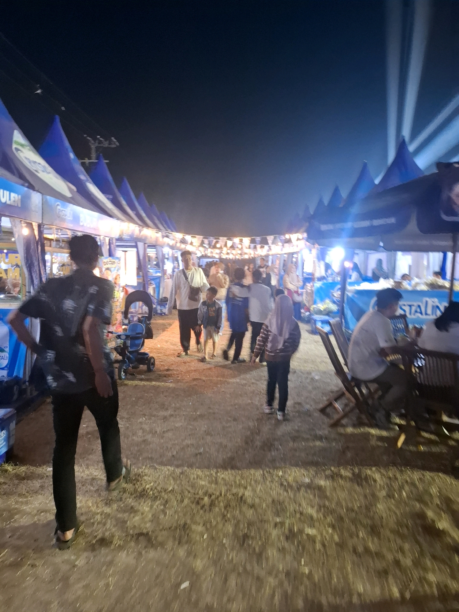 An outdoor night market scene is depicted in Bojong, Indonesia. The foreground shows the lower half of a person from behind, wearing a dark, patterned short-sleeved shirt and black pants. To the left, a blue stroller is visible. In the mid-ground, several individuals are walking. One person in a white shirt and dark pants, holding a bag over their shoulder, is walking with a child in a denim jacket. Further back, a group of people are walking in the same direction. The market stalls, identified by blue triangular awnings, are lit by string lights. In the background, the night sky is dark, with beams of light emanating from the upper right. The ground appears to be dirt and straw.