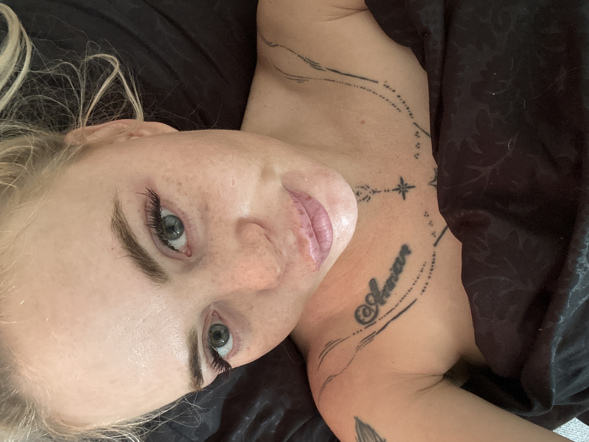 A close-up overhead shot shows a fair-skinned woman with blonde hair and light green eyes. She has numerous freckles across her nose and cheeks, and her eyelashes appear to be enhanced. The woman is lying down, covered by a dark fabric, possibly a blanket or sheets, which has a subtle floral pattern. There are several black tattoos visible on her chest and shoulder area. One tattoo on her chest reads "Amor" in cursive. Another tattoo on her shoulder consists of abstract lines and a starburst design. The background is dark and indistinct, suggesting an indoor setting with low lighting. The woman's expression is relaxed. The image is a selfie, as indicated by the angle and framing.