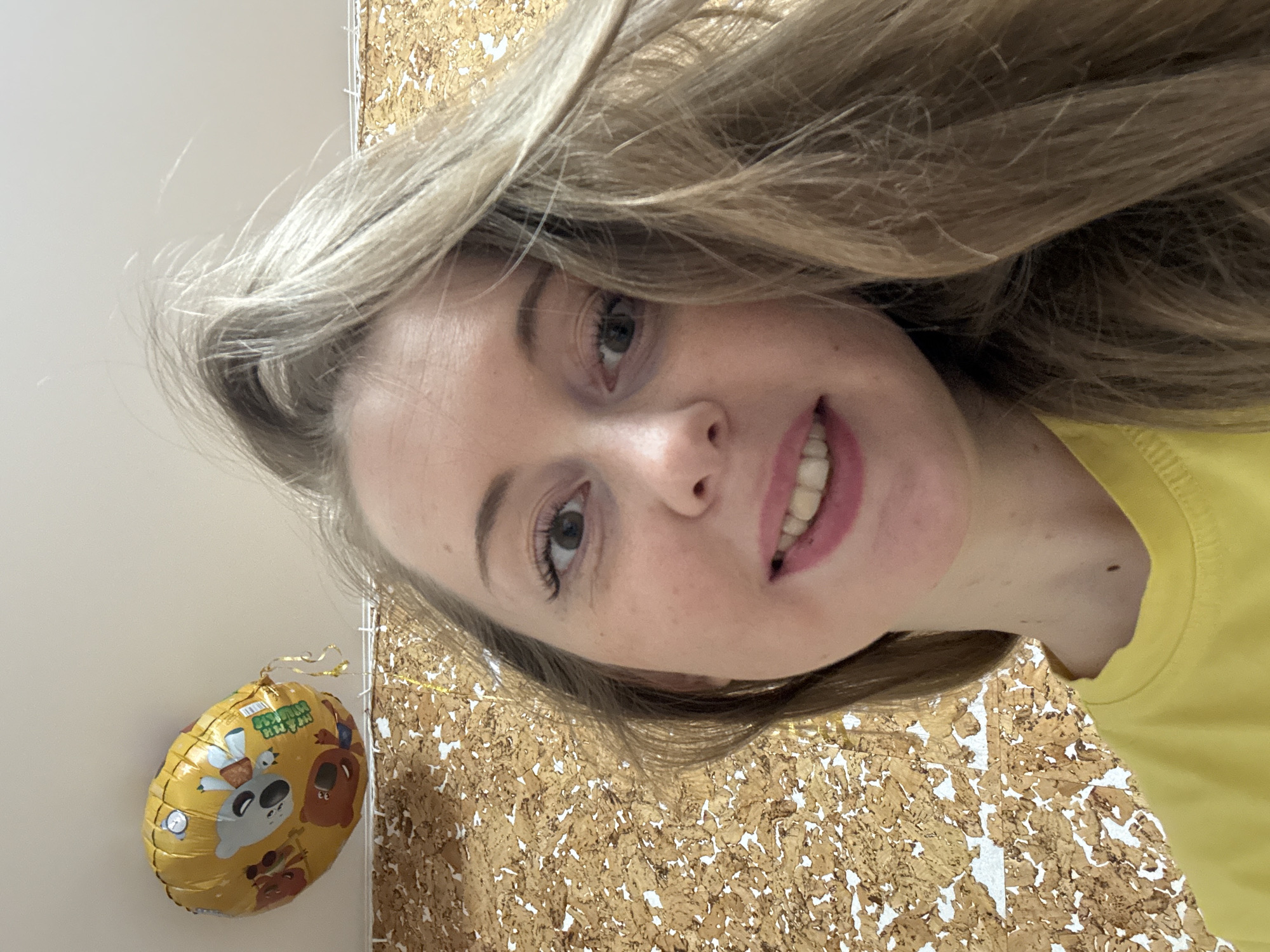 The image is a slightly tilted, close-up selfie of a young woman smiling and looking directly at the camera. She has long, light blonde hair that frames her face and flows over her shoulders. Her skin is fair, and she has light-colored eyes, with visible eyelashes. She is wearing a yellow top, parts of which are visible on her right side. Her lips are parted in a cheerful smile, revealing her teeth.
In the background, on the left side of the image, is a golden, oval-shaped foil balloon. The balloon is decorated with several cartoon animal characters, including a grey koala-like figure, a brown bear, and other smaller animals. A green banner on the balloon clearly displays the English text "HAPPY BIRTHDAY" and below it, Korean characters that read "생일 축하해" (Saengil Chukhahae), also meaning "Happy Birthday." The presence of the balloon suggests a celebration or birthday event.
The setting is indoors. Behind the woman's head and to her right is a distinct wall covering featuring a textured pattern of irregularly shaped golden-brown cork pieces interspersed with white spaces, creating a mosaic or distressed effect. To her left, behind the balloon, is a plain, light-colored wall, possibly white or off-white. The lighting appears bright and even, indicating daytime conditions indoors. The woman's smile conveys a happy and cheerful emotion.