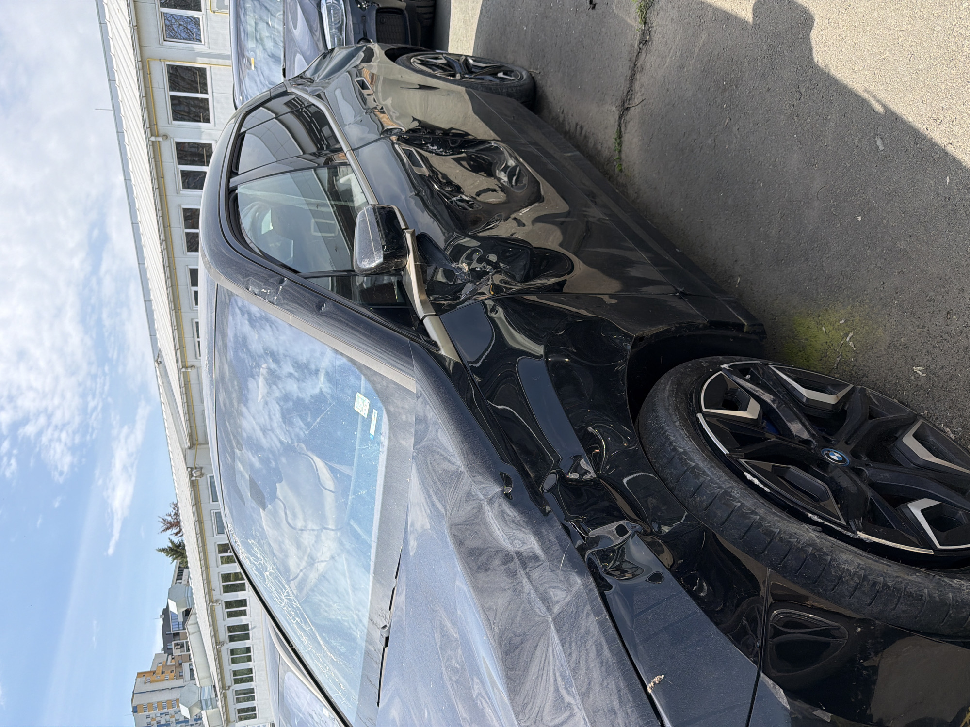 The image displays a black car, identified as a BMW by the logo on its wheel, severely damaged along its entire left side. The front fender, front door, and rear door are extensively dented and crumpled, indicating a significant collision. The front left wheel's rim also shows visible scrapes and damage.
The car is stationary, parked outdoors on an asphalt surface, which appears to be a street or parking lot. To its left stands a large building with a white corrugated roof and numerous windows, suggesting an office or industrial complex. Another vehicle is partially visible in the background behind the damaged car.
The scene is illuminated by bright daylight under a clear sky with scattered clouds, casting sharp shadows on the ground. This suggests the photograph was taken during the mid-day or afternoon. The car's windshield has reflections of the sky and building, along with some visible dust or smudges. A human-like shadow is cast on the ground near the front of the car, though no people are directly visible. The only clear text visible is the "BMW" logo on the center cap of the front left wheel.