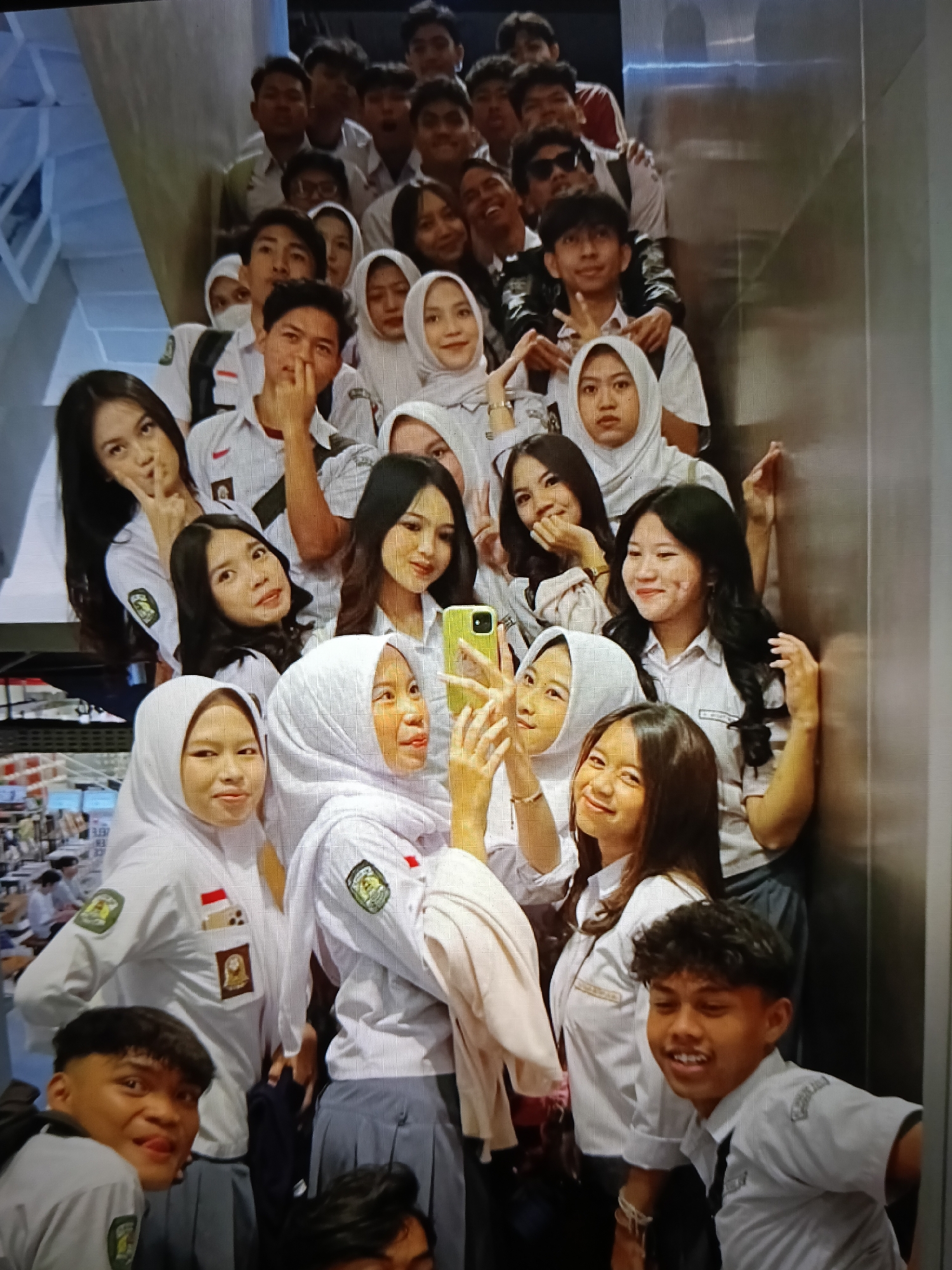 A group of Indonesian students, mostly teenagers, are posing for a photo on a staircase within what appears to be a school or public building in Raha, Indonesia. The students are wearing white uniforms, many with red and green accents, and some are wearing headscarves. They are positioned on multiple steps of a wide staircase, with some in the front row and others behind them. Several students are looking at the camera, with varying expressions ranging from smiles to playful poses. One student in the front row is holding a yellow smartphone, suggesting they might be taking a selfie or documenting the moment. The environment appears to be indoors, with a metallic-looking wall to the right of the staircase. The lighting is somewhat diffused, casting shadows and highlighting the faces of the students.