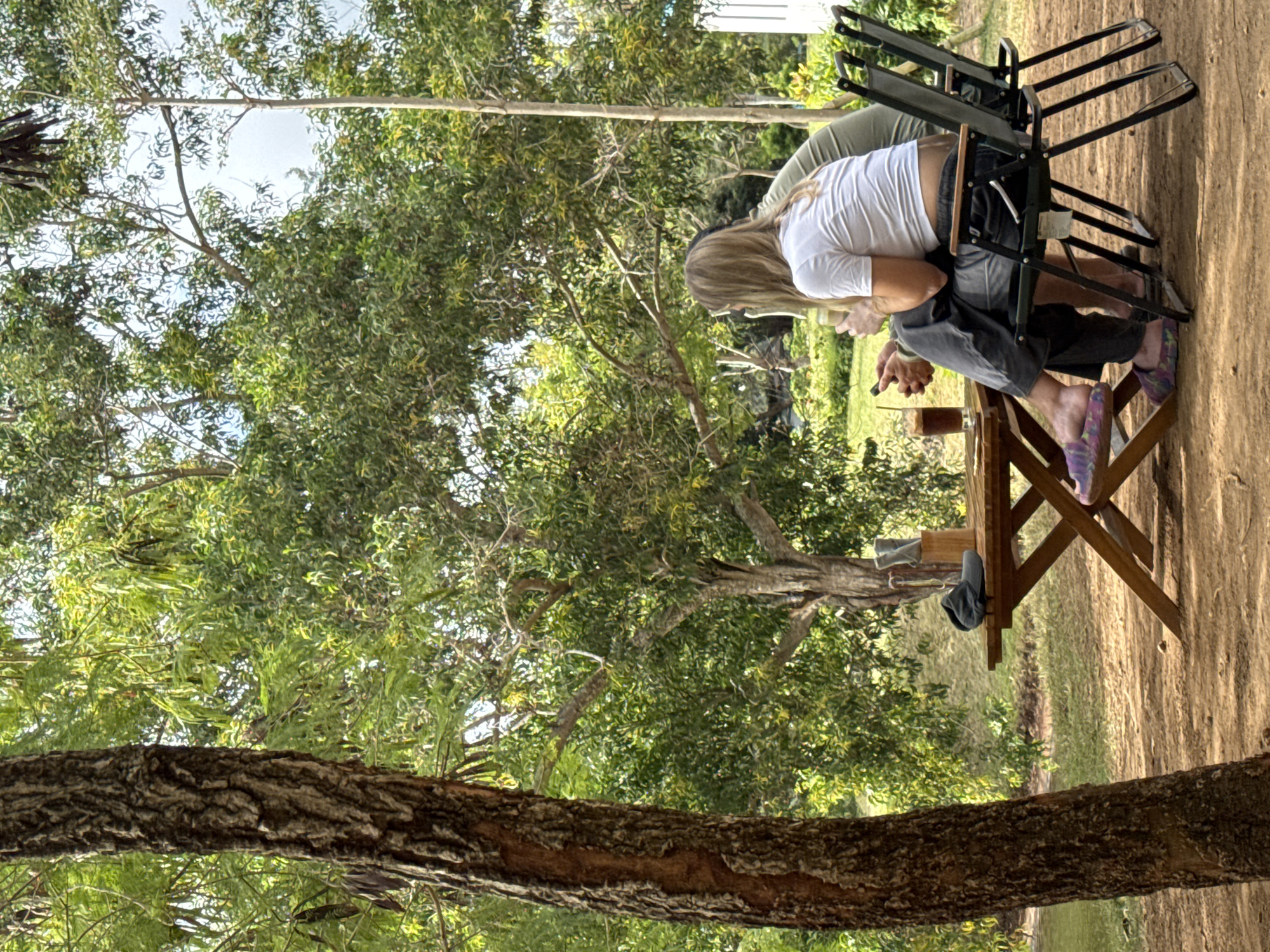In Kuta Selatan, Indonesia, two individuals are seated at a wooden table outdoors, beneath the canopy of leafy trees. The person on the left, facing away from the camera, has long blonde hair and wears a white t-shirt and dark pants. They are holding a drink with a straw and a mobile phone. The person to their right, partially visible, wears a green shirt. On the table, a glass with a dark beverage and a straw, along with a hat and what appears to be a napkin holder, are present. Two foldable chairs are positioned at the table. The ground is covered in reddish-brown dirt, and the background features lush green foliage and trees.