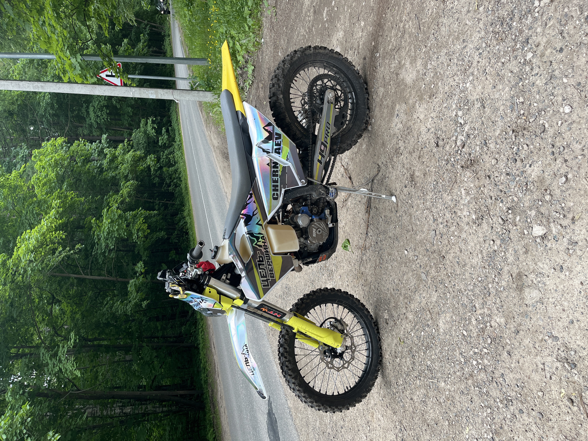 A dirt bike, primarily white and yellow with holographic decals, is parked on a gravel and dirt shoulder beside an asphalt road in Ореховка, Russia. The motorcycle is equipped with knobby tires, disk brakes, and a visible clear plastic fuel tank. Decals on the front fender and tank shrouds display "ЦЕЛЬ ВЕРШИНА" (Target Summit) with mountain graphics. The right side panel near the seat shows "CHERNAEV," and the swingarm displays "H9PRO." The front forks and rear fender are yellow. The bike is resting on its extended kickstand, facing left.
To the left of the road, a dense green forest with tall deciduous trees dominates the background. The asphalt road features a white dashed center line and curves gently to the right in the distance. On the right side of the road, a grassy area with small yellow flowers is visible. A concrete utility pole and a metal pole stand upright on the right side of the road, near a red and white triangular road sign indicating a T-intersection where a side road joins from the right, with the main road curving left. A dark-colored vehicle is partially visible on the road in the far distance to the right. The scene is illuminated by natural daylight, suggesting an overcast or partly cloudy sky. No individuals are visible in the image.