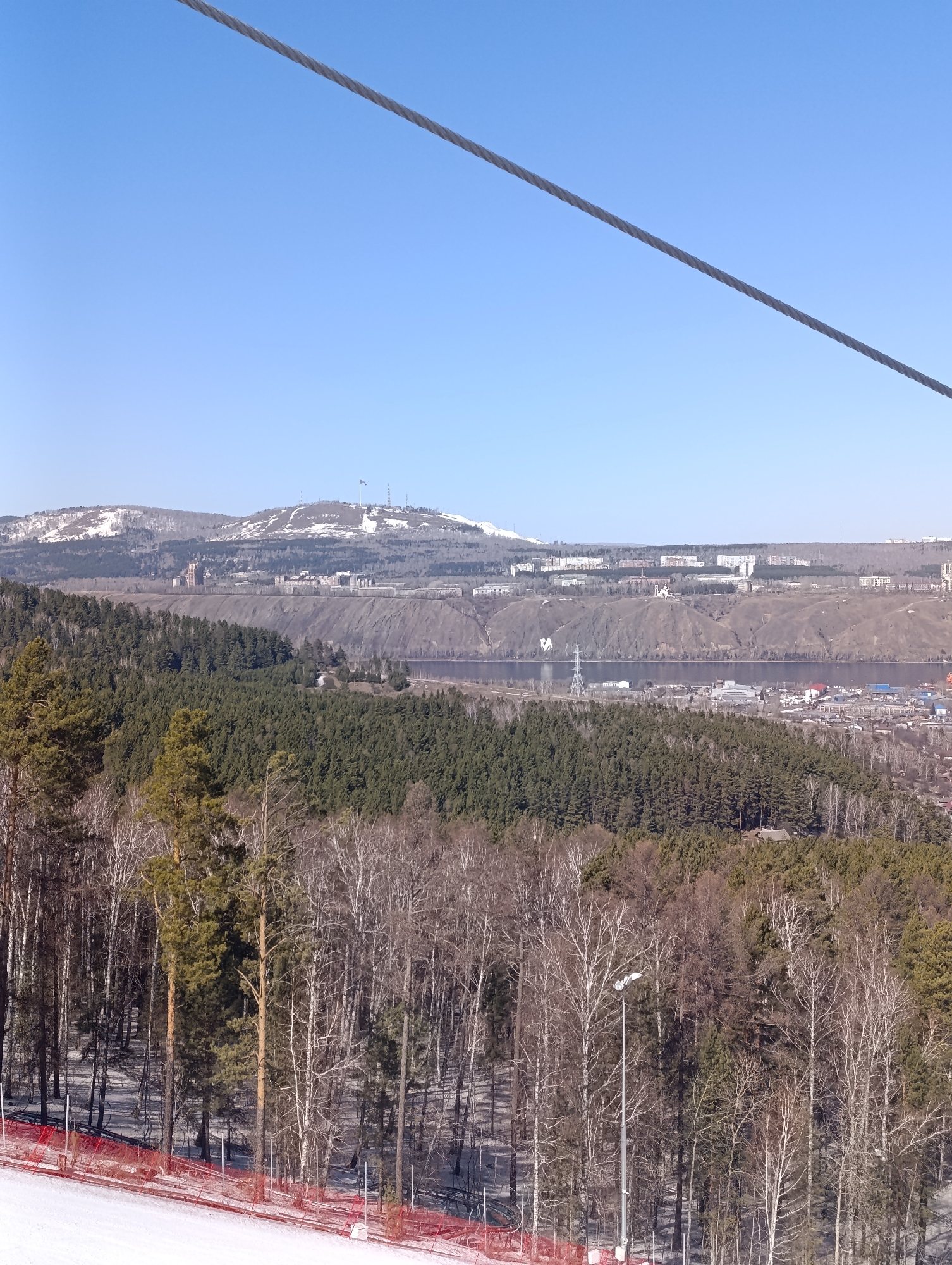 The image captures a scenic view from a ski slope in Krasnoyarsk, Russia. The foreground features a snow-covered slope with red safety netting. Behind the slope, a dense forest transitions from coniferous to deciduous trees. A street lamp is visible among the trees. In the distance, the Yenisey River flows, separating a populated area with buildings and infrastructure from a mountain range. The mountain range has snow-capped peaks and structures, including communication towers, on top. A cable, possibly part of the ski lift infrastructure, runs diagonally across the top of the image. The sky is clear and blue.