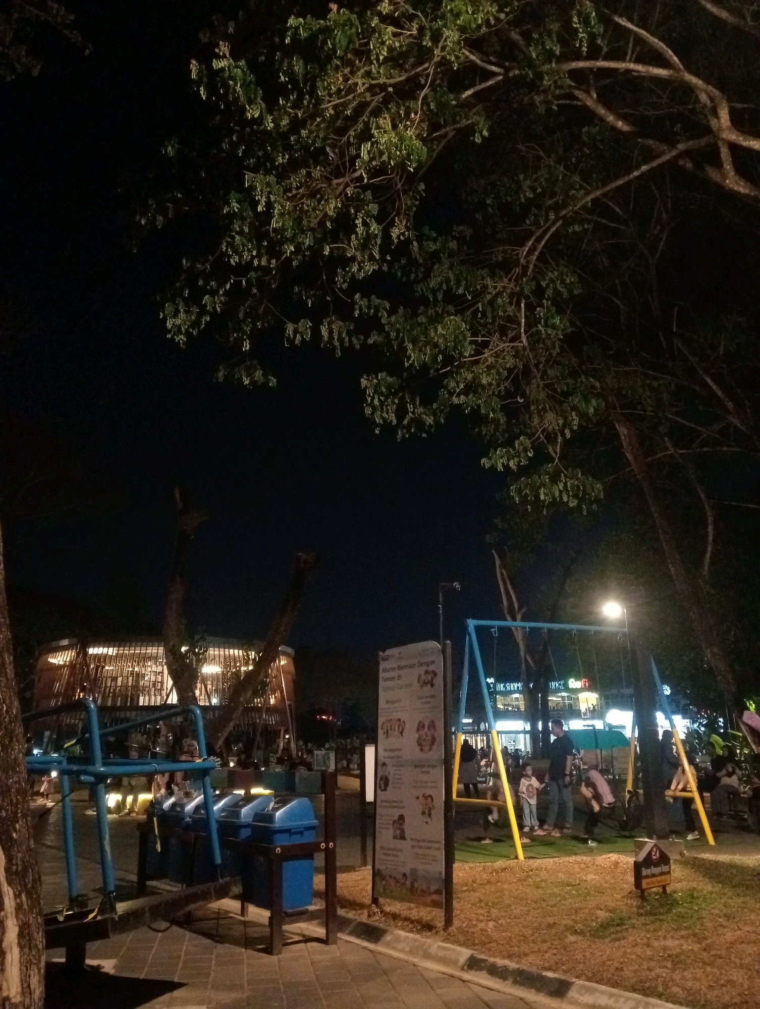 This is a nighttime outdoor scene at a playground in Samarinda, Indonesia, possibly along the Mahakam River waterfront, given the general ambiance and presence of a modern building in the background that could be a commercial establishment or a well-lit public area.
In the foreground, a blue metal swing set is prominent, with several children and adults visible. One child in a light-colored shirt is on a swing, while others stand nearby. A man in dark clothing is observing the children. To the left of the swing set, a row of blue trash bins is situated. A signpost with some Indonesian text and illustrations is also visible to the left, likely containing playground rules or information.
The background features a modern, circular building with warm interior lighting, suggesting it might be a restaurant, cafe, or shop. The overall environment is illuminated by artificial lighting, casting shadows and highlighting the trees overhead. The ground appears to be a mix of paved walkways and grassy or sandy areas. The presence of mature trees with large branches adds to the park-like setting. The atmosphere is casual, with people enjoying the evening in a public recreational space.