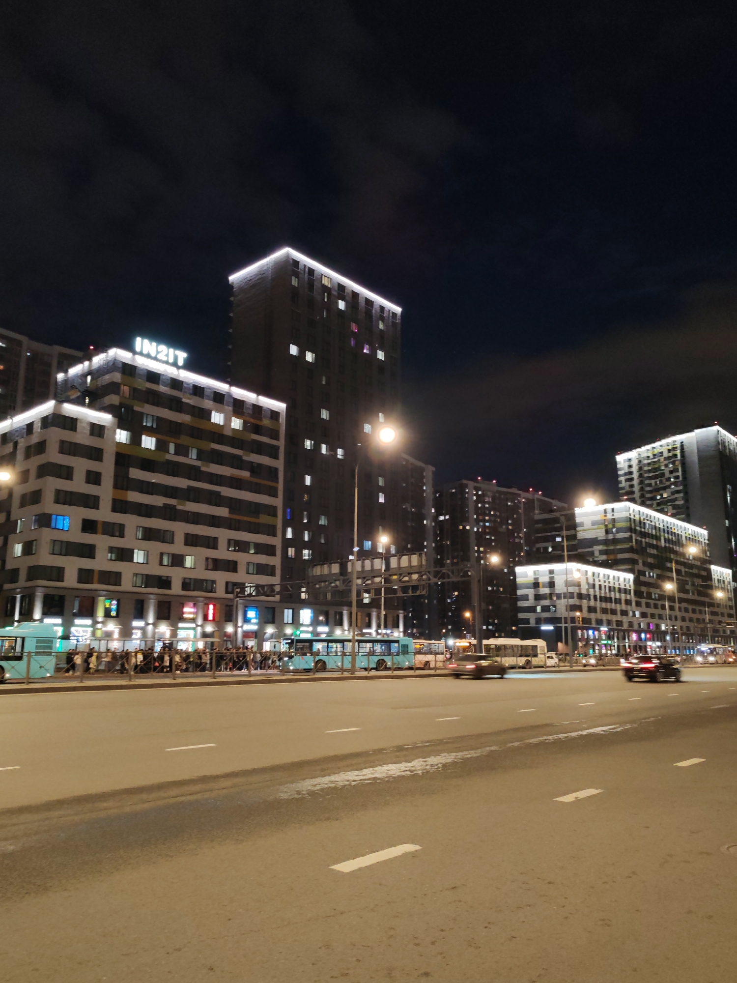 The image captures a dynamic urban street scene at night in Saint Petersburg, Russia. Modern, multi-story buildings with distinct architectural features and numerous illuminated windows dominate the background. Many of these buildings are adorned with decorative white light strips along their upper edges, creating a bright skyline. The most prominent building on the left proudly displays a large, white, illuminated sign with the text "IN2IT."
In the foreground, a wide, multi-lane asphalt road stretches into the distance, marked with white dashed lane lines. Numerous vehicles are present, including cars and several light blue public buses, as well as a white bus. Some cars are captured with motion blur, indicating active traffic on the road.
On the far side of the road, a bustling sidewalk is visible, populated by a significant gathering of people. Many individuals are standing near the light blue buses, suggesting a bus stop or public transportation hub. Streetlights punctuate the scene, casting bright pools of light that complement the illumination emanating from the buildings and vehicle headlights and taillights.
The sky is dark, indicating nighttime, with faint cloud formations visible against the deep blue-black expanse. The overall atmosphere is one of a vibrant and active city evening, characterized by modern architecture and ongoing urban life.