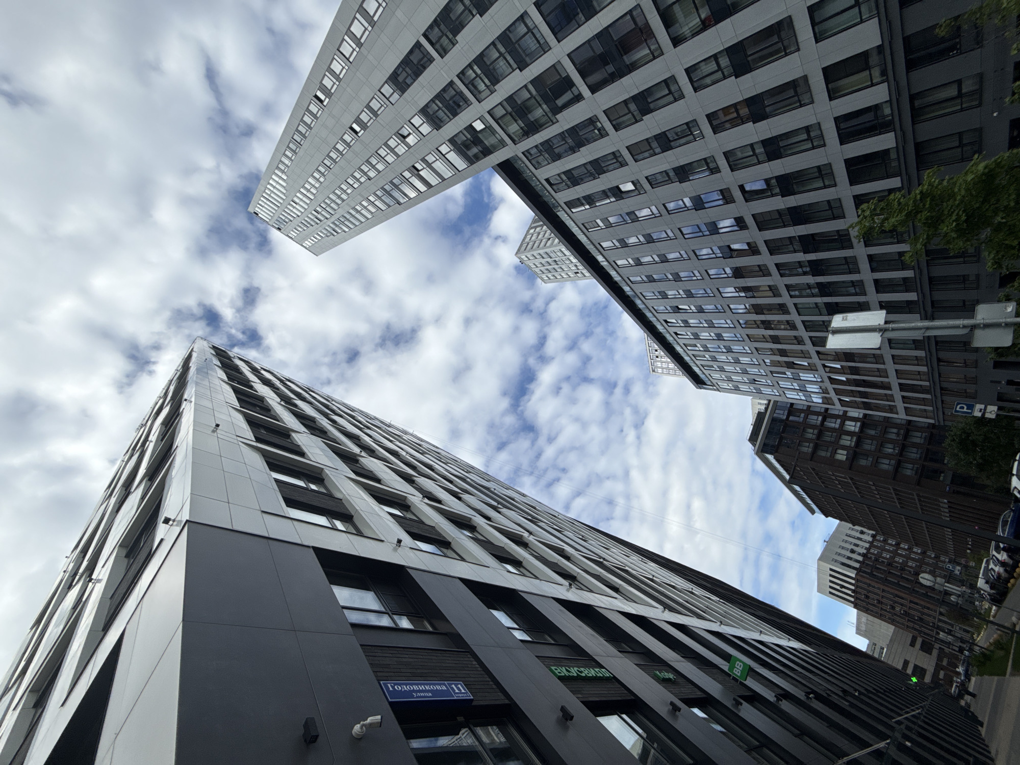 The image shows a low-angle view of modern architecture in Moscow, Russia. Two high-rise buildings with a predominantly gray and white facade are visible against a sky filled with scattered clouds. The building in the lower part of the image is partly clad in dark gray and has signage in Russian, including a street sign for "Godovikova 11". Other buildings, including one with a brown facade and visible windows, are visible in the background. A street scene with parked cars can be seen on the right side of the image, with a few green trees. No people are visible in the shot.