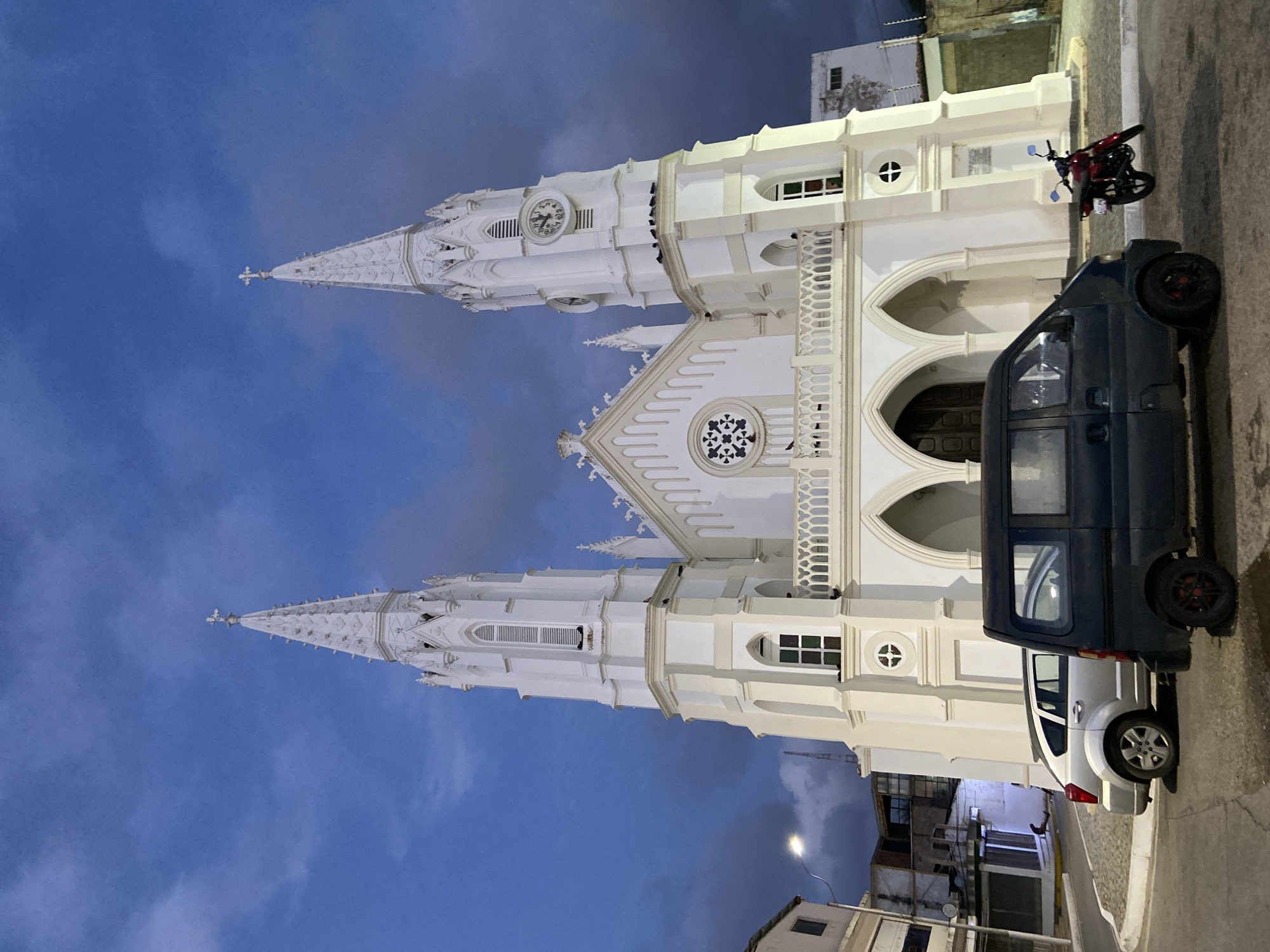 The image depicts a white church with two spires, a clock on one of the spires, and ornate architectural details. The sky is a deep blue with dramatic clouds, suggesting it might be dusk or dawn. In front of the church, there are two vehicles: a silver car and a dark van. A red motorcycle is parked to the right of the van. In the background, to the left of the church, are some buildings. The scene appears to be in an urban or semi-urban setting, possibly a town square, given the presence of vehicles and buildings surrounding the church. The church is likely the Iglesia de San Juan Bautista in Juan Griego, Venezuela, known for its distinctive twin spires and prominent clock. The lighting and overall atmosphere suggest a quiet moment in the town.