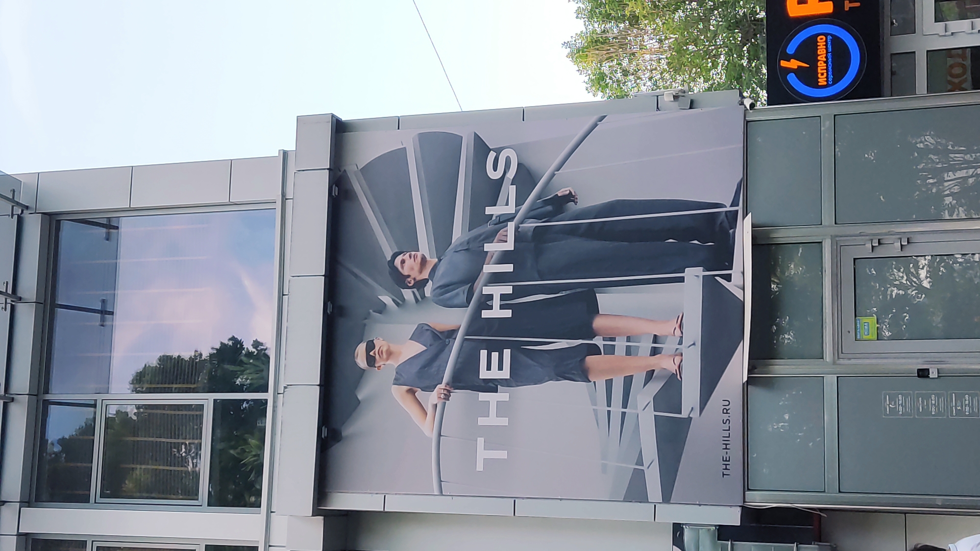 This is a medium-shot, eye-level, exterior photograph of a commercial building with a large advertisement banner. The banner features two models, a woman in the foreground and a man behind her, both dressed in dark clothing and posing on a spiral staircase. The text "THE HILLS" is prominently displayed on the banner, along with "THE-HILLS.RU". The building has large windows reflecting the sky and trees. Below the banner, there are storefront windows and doors. To the right of the banner, a neon sign is visible with Cyrillic text that reads "ИСПРАВНО" within a blue circle and lightning bolt symbol, suggesting a repair service. This building is located in Krasnodar, Russia, specifically on Uritskogo Street, 220.