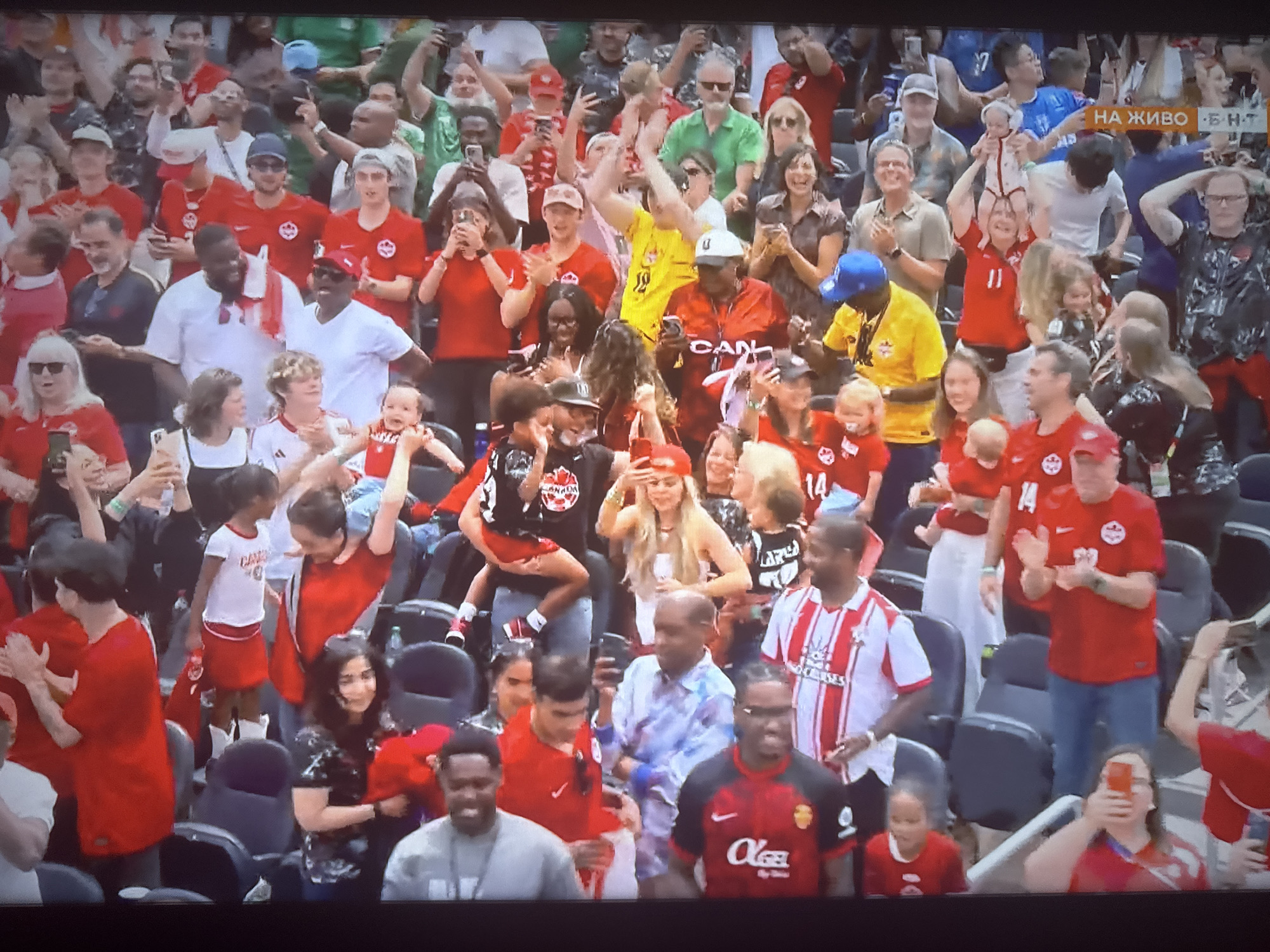 The image shows a crowd of people at an outdoor event, likely a sports game given the jerseys and fan attire. The crowd is predominantly dressed in red, many wearing jerseys with Canadian iconography. Several individuals are holding up phones, presumably to take pictures or videos. Some people are clapping, suggesting excitement or engagement with the event. The background is filled with spectators seated in stadium-like seating. The lighting suggests it's daytime. The environment appears to be a large public gathering space with a focus on the event taking place.