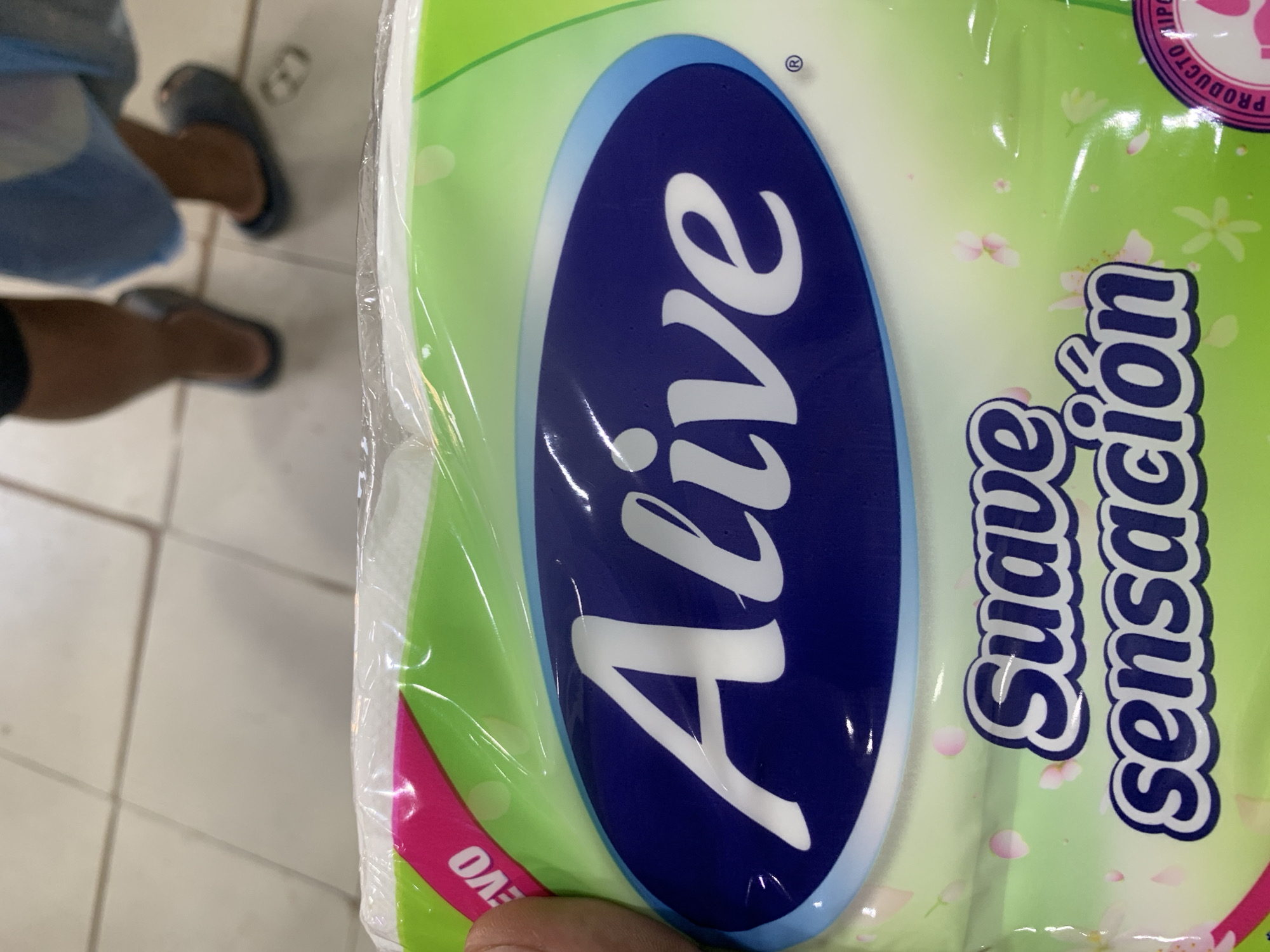 A close-up, eye-level shot captures a package of "Alive" brand toilet paper, possibly in a commercial or retail setting in Santa Ana, Venezuela. The packaging features a bright green background adorned with scattered pink flowers and petals, evoking a sense of freshness and delicacy. Prominently displayed on the package is the brand name "Alive" in a stylized white font within a dark blue oval. Below this, in smaller white lettering with a dark blue outline, are the words "Suave sensación," which translate to "Soft sensation." A small pink tag on the upper left corner reads "NUEVO" (New). The packaging appears to be wrapped in clear plastic, reflecting light and creating glare. In the blurred background, two pairs of feet are visible, clad in dark slippers, suggesting the presence of shoppers or employees. The floor appears to be tiled in a light, neutral color. The overall impression is of a product displayed for sale, highlighting its sensory qualities.