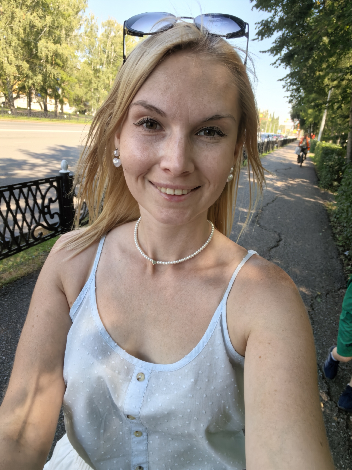 In Sterlitamak, Russia, a woman with blonde hair and pearl jewelry takes a selfie outdoors on a sunny day. She is wearing a light blue, sleeveless top with small white polka dots and buttons down the front. She has sunglasses perched on top of her head. She is smiling and looking at the camera. To her left, a black decorative fence lines a sidewalk. To her right, a paved path runs alongside a hedge and trees, with a person on a bicycle visible in the background. A child wearing green pants and dark shoes is partially visible on the far right of the image.
