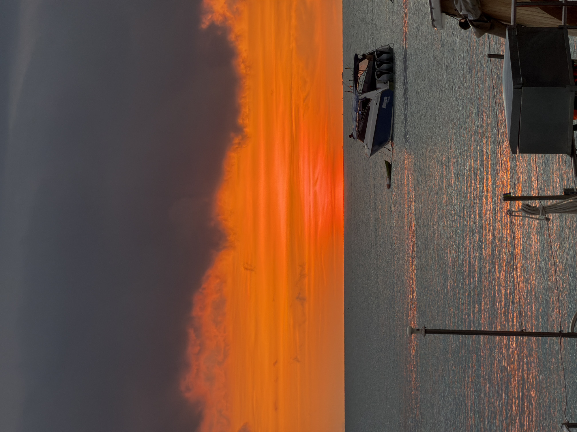 An image taken at sunset in Baan Bang Rak, Thailand, features a dramatic sky with dark gray clouds above vibrant orange and red clouds. The sun is setting on the horizon, casting a warm glow on the water. A speed boat with a blue and white hull is anchored in the calm sea, accompanied by a small yellow dinghy. In the foreground, parts of another boat are visible, including a metal box and a pole with a light fixture. The water reflects the colors of the sky, creating a shimmering effect. The scene is peaceful and serene, capturing the beauty of a tropical sunset.