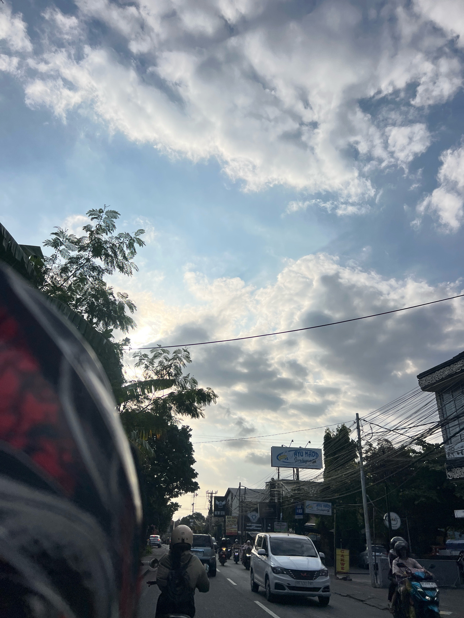 The image captures a street scene in Denpasar, Indonesia, under a cloudy sky with visible sunlight filtering through. A person is riding a motorcycle in the foreground, partially obscuring the view with their helmet. Other vehicles, including cars and motorcycles, are present on the road, indicating moderate traffic. Buildings and trees line the street, with utility wires prominently displayed above. A billboard with the text "AYU NADH Girelaysan" is visible on one of the buildings. The overall atmosphere suggests a typical day in a bustling urban environment.