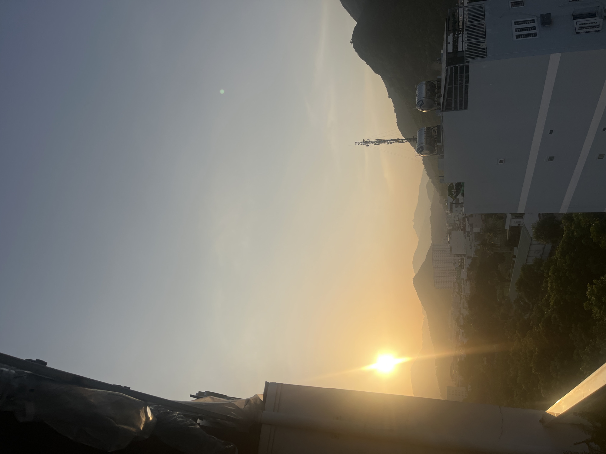 The image shows a sunset over the city of Nha Trang, Vietnam. The sun is low on the horizon, casting a warm glow over the landscape. In the foreground, there is a building with a white facade and a balcony. In the background, there are mountains and a cityscape with buildings and trees. The sky is clear and blue, with a few wispy clouds. The overall mood of the image is peaceful and serene.