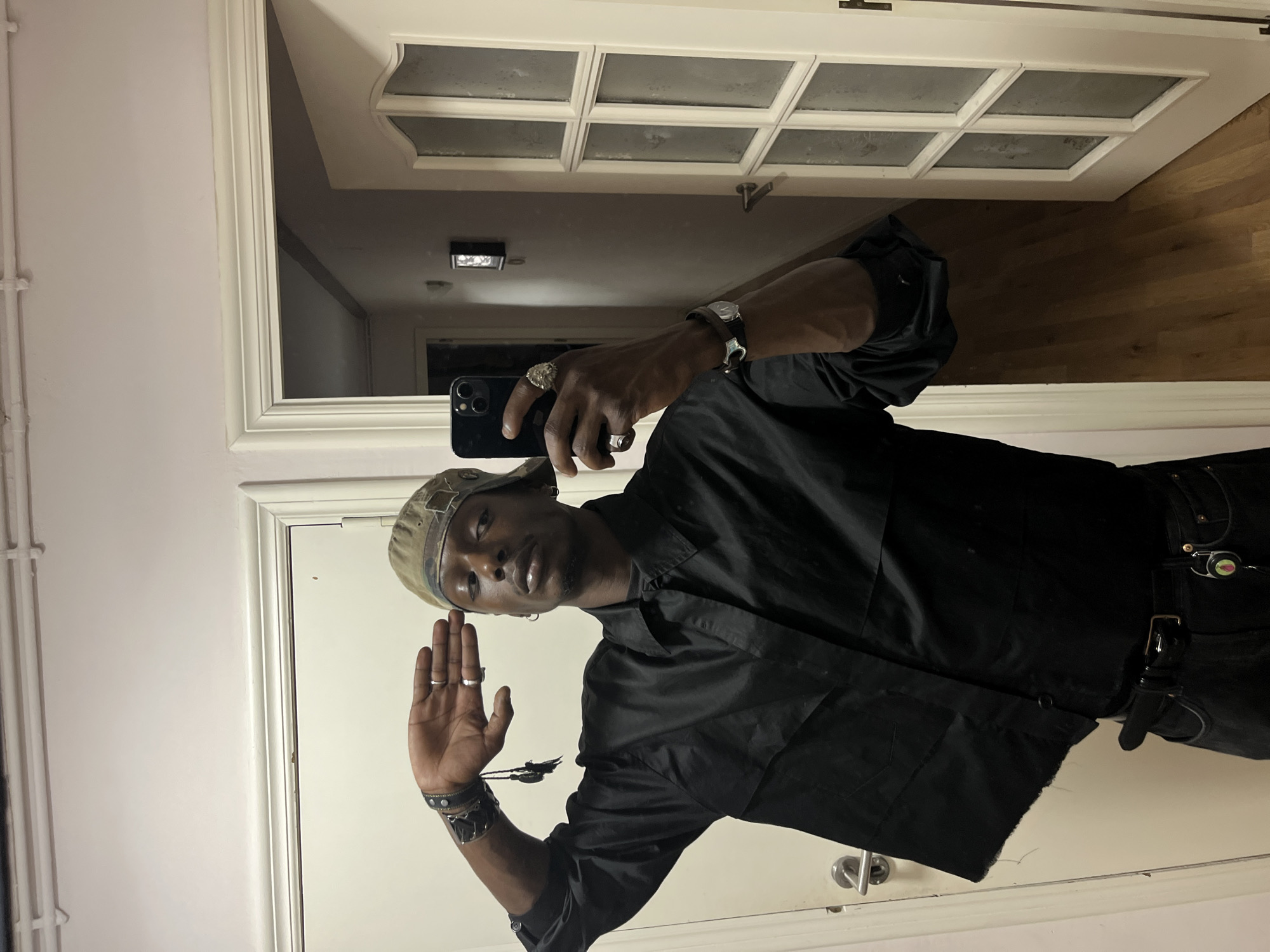A male subject takes a mirror selfie indoors, rotated 90 degrees clockwise. He has a dark skin tone and is wearing a black long-sleeved collared shirt, black pants with a prominent black belt, and a camouflage-patterned bucket hat. He holds a black smartphone (appearing to be an iPhone with multiple cameras) in his right hand, angled towards his reflection. His left hand is raised with the palm open and fingers slightly splayed.
The man wears several silver rings on his fingers, a watch, and multiple black leather bracelets or cuffs, some with studs, on his left wrist. A small, colorful trinket or charm is attached to his belt. He looks directly at the camera with a neutral expression.
The background, seen through the mirror's reflection, reveals a light-colored room. To the left of the mirror, a white wall with white piping (possibly a radiator) is visible. The mirror itself is framed in white. Reflected above, a white panelled door with multiple square glass inserts is visible, along with a white ceiling featuring a square recessed light fixture. A light-colored wooden floor is reflected at the bottom of the mirror. A metallic door handle is also visible on the white door frame. The setting appears to be an interior space, such as a hallway or apartment in Barcelona, Spain, illuminated by artificial light. No visible text is present.
