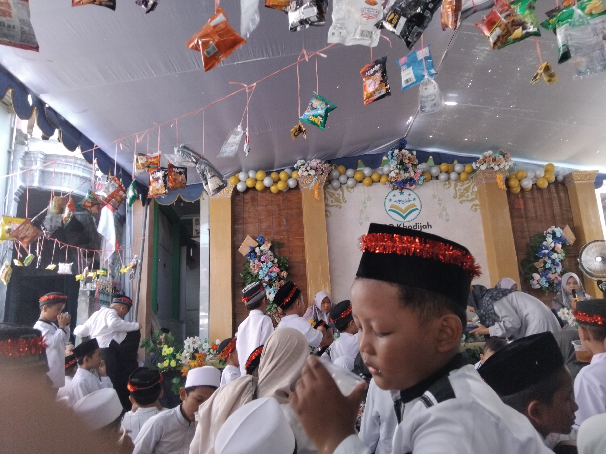 In Kepuh Kiriman, Indonesia, a group of children and adults are gathered under a white tent structure. Bags of snacks are hung from strings across the tent, creating a festive atmosphere. Several children are wearing white shirts and black caps, some with red trim. Some children appear to be holding cups or drinks. In the background, there is a wall decorated with balloons and flowers, as well as a banner with Arabic calligraphy. The overall scene suggests a celebration or event.
