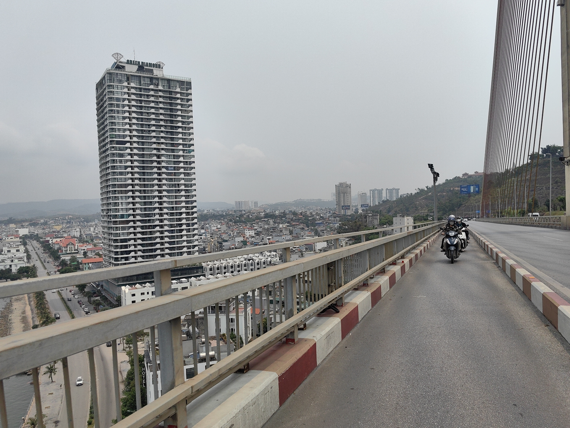The photo shows a view from a bridge in Hong Gai Ward, Vietnam. In the background, a tall building labeled "GREEN DIAMOND" dominates the skyline, surrounded by cityscape and hills. In the foreground, two motorcycles are traveling on the road next to a pedestrian railing. The sky is overcast.
