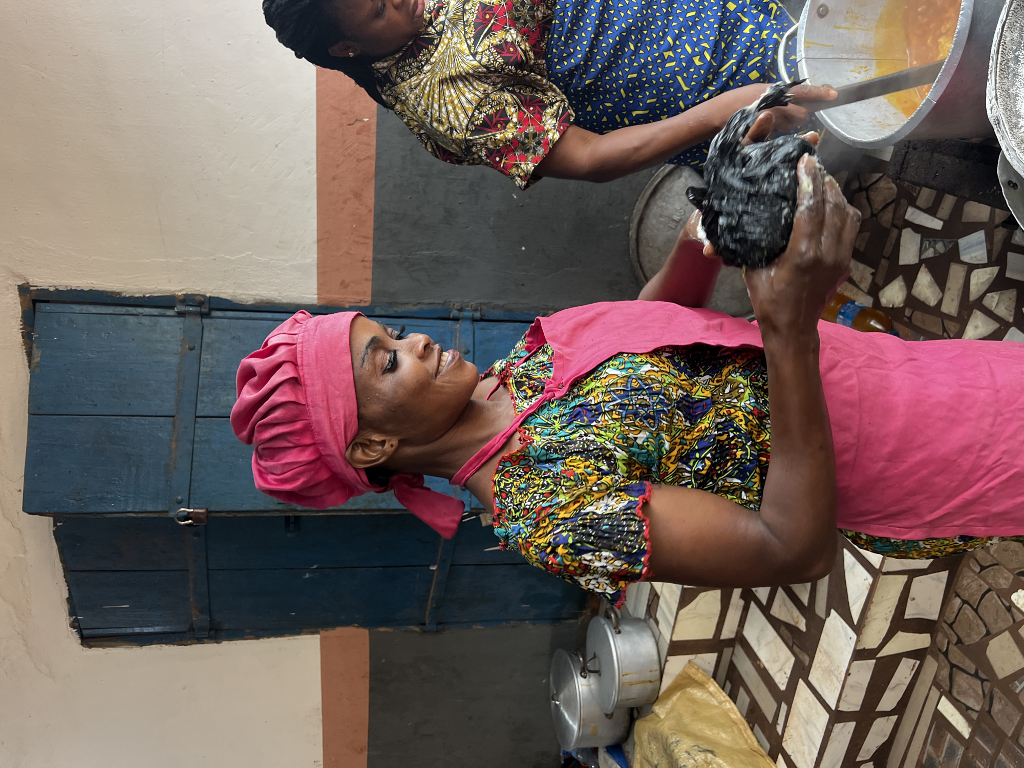 In Nkpor, Nigeria, two women are engaged in a food preparation activity. The woman in the foreground, wearing a pink headscarf and apron over a vibrantly patterned dress, smiles as she holds a dark, lumpy object, possibly a protein source like meat or fish, which she is cleaning or preparing. Her expression suggests a sense of satisfaction or focus. Behind her, another woman, in a blue and yellow patterned dress, is partially visible, leaning over a large pot. She appears to be stirring or attending to something in the pot, which contains a yellow, oily liquid, likely cooking oil.
The scene unfolds in an indoor setting with a rustic feel. To the left, a blue wooden door with metal latches is prominent, suggesting a shop or a traditional dwelling. The floor is covered with a mosaic of irregularly shaped tiles in beige and brown hues. Several metal pots are visible, indicating a kitchen or food stall environment. A yellow bag lies on the floor near the pots. The lighting suggests it could be daytime, possibly mid-morning or afternoon, as there are no strong shadows indicating early or late sun. The overall atmosphere is one of work and community, with the women focused on their task of preparing food.