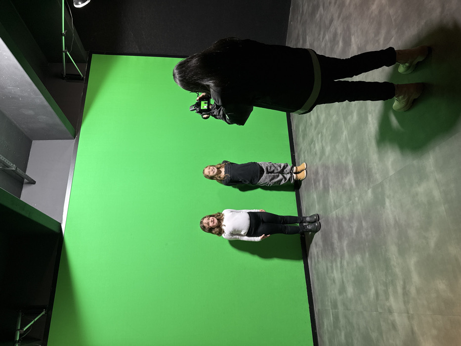 This image depicts an indoor photography session in a studio located in Canela, Brazil. Two children, appearing pre-adolescent, stand side-by-side facing forward in front of a large, illuminated green screen that extends from floor to ceiling. The child on the left wears a white long-sleeved shirt, black pants, and dark boots. The child on the right wears a dark long-sleeved shirt, grey patterned cargo pants, and light brown boots. Both children are smiling faintly. In the foreground, an adult, viewed from the back, holds a black DSLR camera aimed at the children. This adult wears a dark jacket with a white band at the lower hem, dark pants, and light-colored boots. The studio environment features dark, possibly black, walls and ceiling, with visible dark structural elements and pipes overhead. The floor is composed of large, mottled grey square tiles. Shadows from the subjects are cast onto the tiled floor directly in front of the green screen.