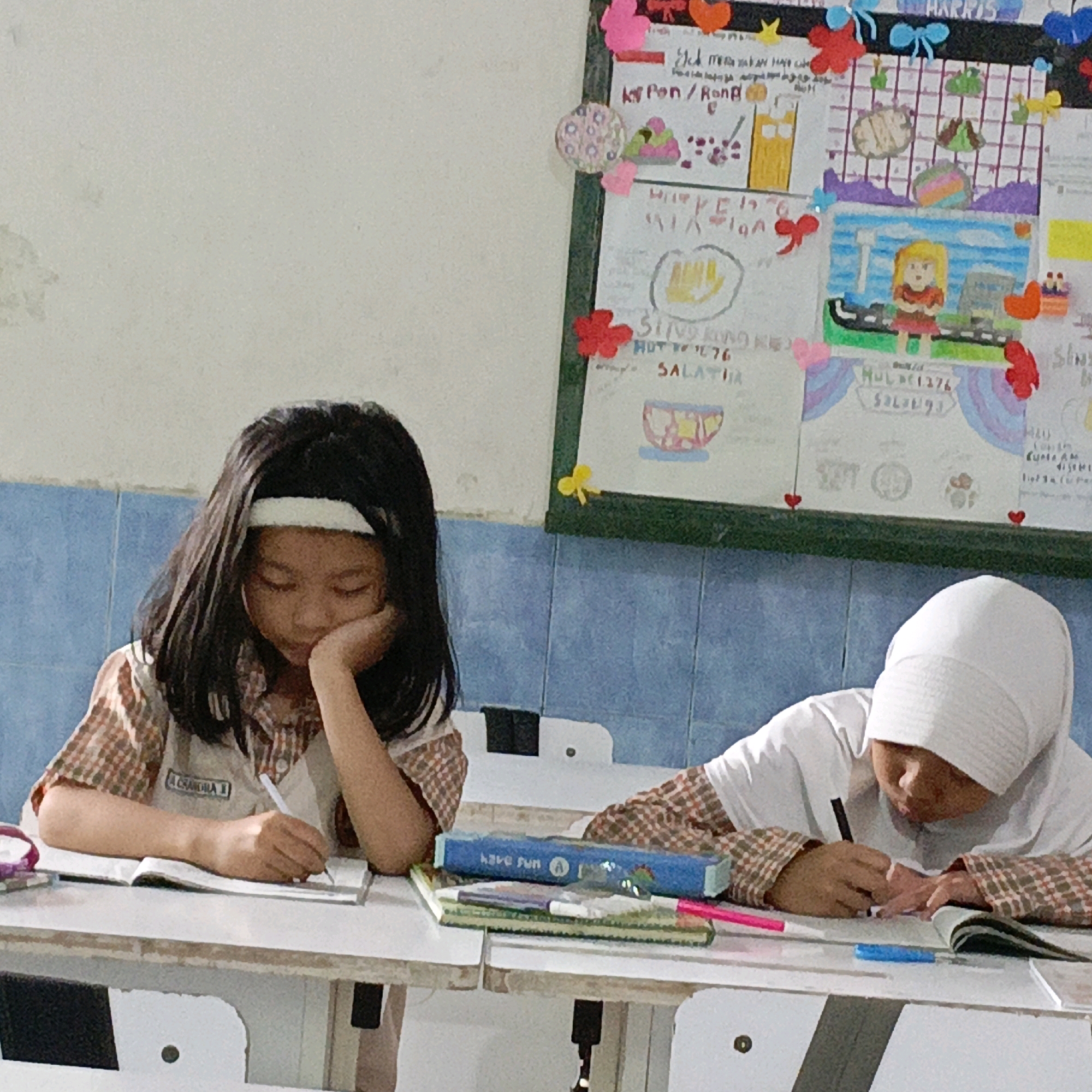 In a classroom in Salatiga, Indonesia, two young girls are engaged in academic activities. On the left, a girl with long dark hair and a white headband rests her head on her hand, looking down at her open notebook on the desk. She is wearing a plaid collared shirt. To her right, a girl wearing a white hijab is focused on her work, writing in a notebook with a black marker. Her plaid long-sleeved shirt is visible beneath her white outer garment. Their desks are cluttered with school supplies, including notebooks, pens, and markers. In the background, a colorful bulletin board adorned with drawings, cutouts, and text hangs on the wall. The wall behind them is tiled with blue and white squares.