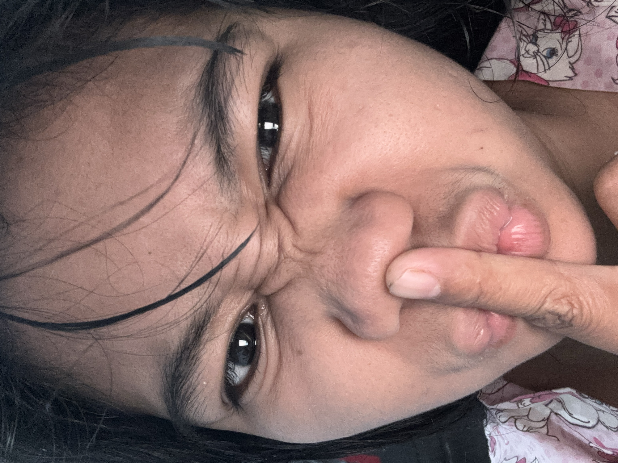 A close-up, high-angle indoor shot captures a young Indonesian woman's face in North Jakarta, Indonesia. Her eyes are narrowed and scrunched, with wrinkles forming around them. Her eyebrows are bushy and dark. A finger is held to her nose, and her lips are pursed in a duck-face expression. Her skin is a light brown, and a few strands of dark hair fall across her forehead. She is wearing a pink shirt with a cartoon print. The background is blurred and dark, suggesting an indoor setting.