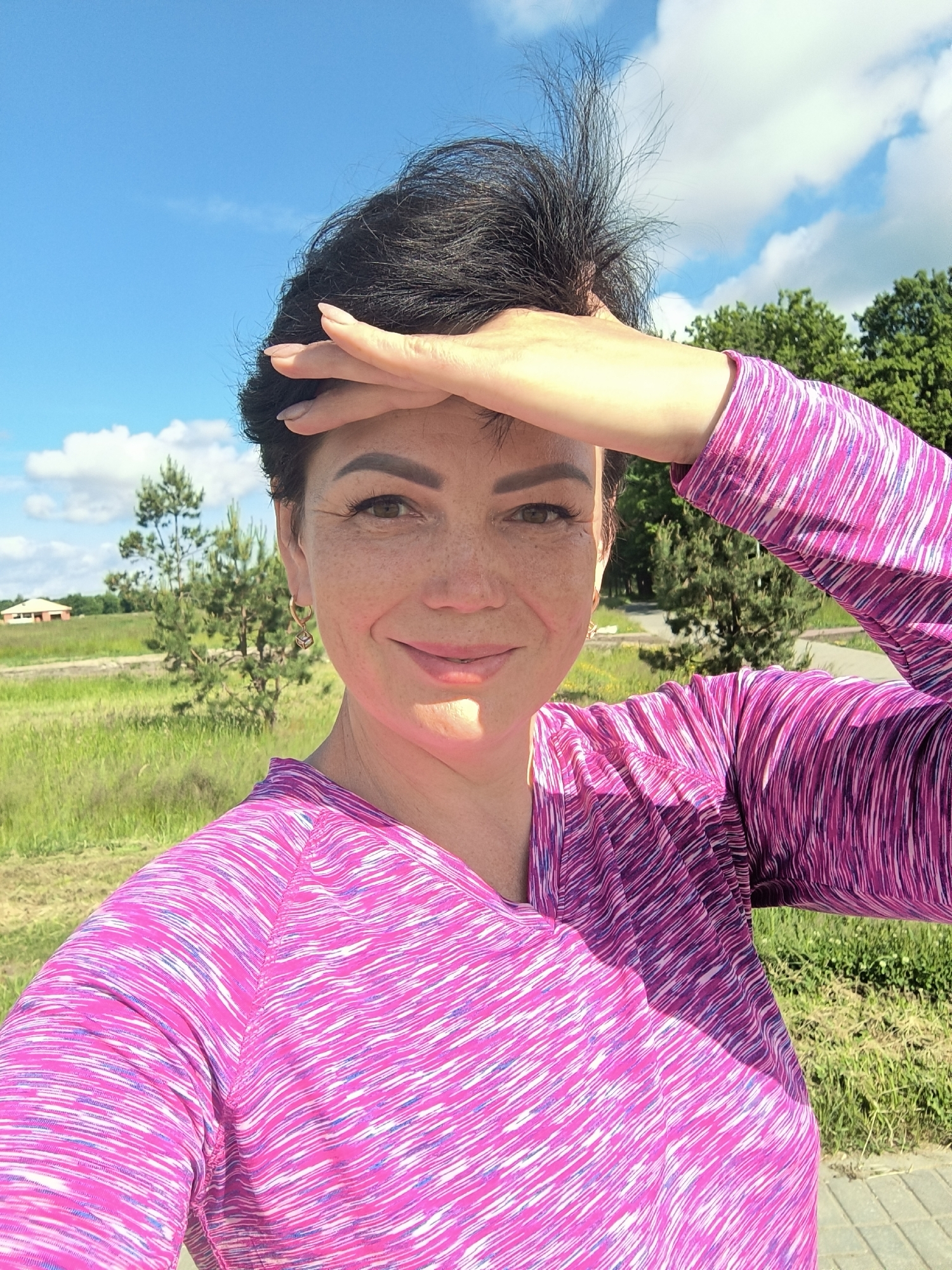 A woman with short, dark hair and freckles is taking a selfie outdoors on a sunny day in Laskino, Russia. She is wearing a pink, long-sleeved shirt with a white and purple striped pattern. Her right arm is raised, and her hand is shielding her eyes from the sun. She is smiling at the camera. The background shows a grassy field with some trees and a blue sky with white clouds. In the distance, a small house is visible.