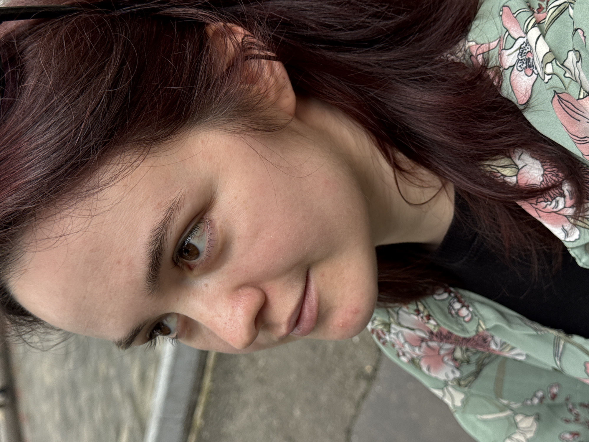 A close-up of a woman's face and upper body. She has auburn hair and fair skin. She is wearing a floral-patterned shirt. The background is indistinct, with hints of grey possibly indicating a concrete surface. There is no evidence of Sochi, Russia.