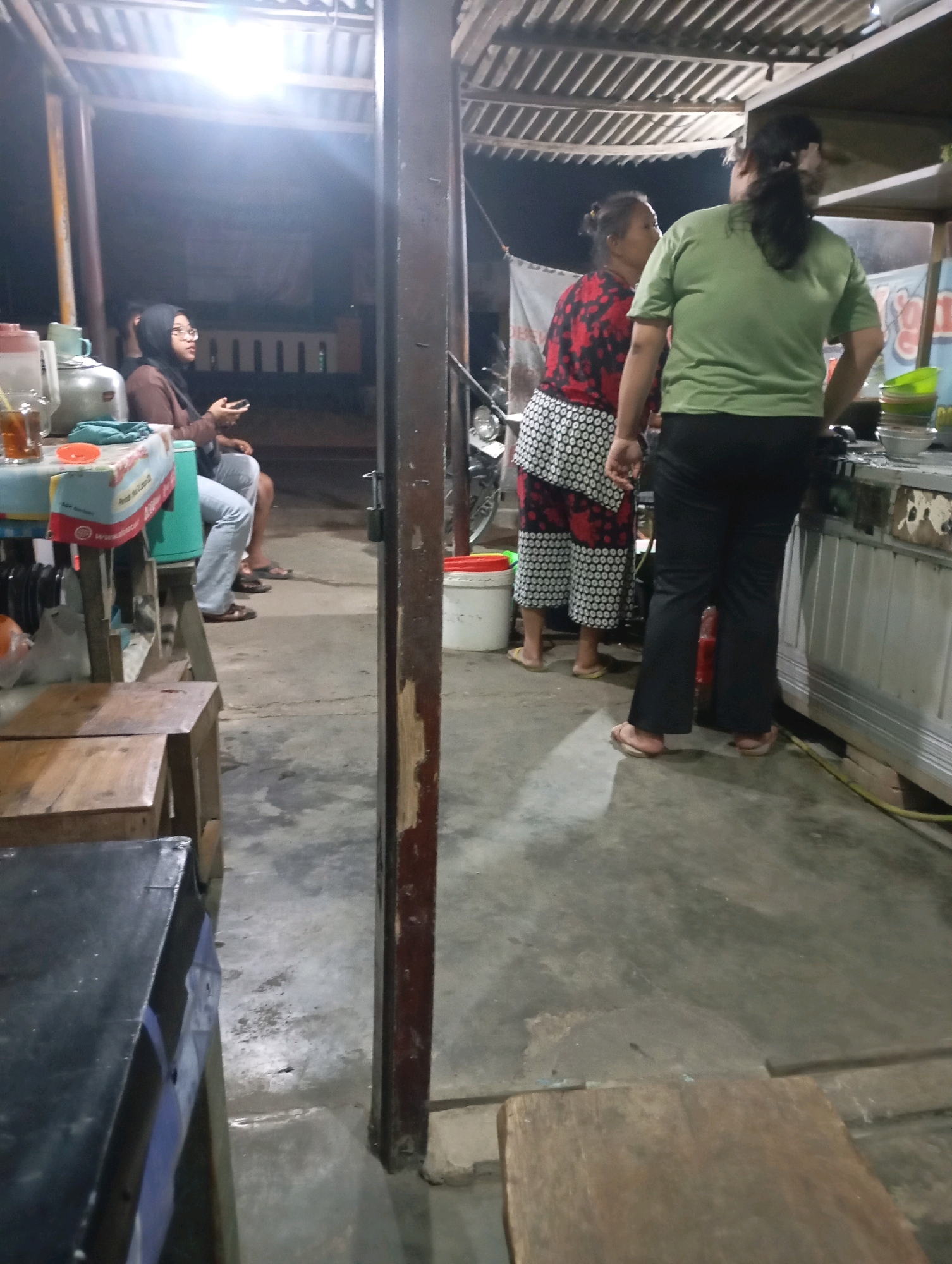 This is a medium shot, taken at night, of a food stall in Pacitan, Indonesia. Three people are present. In the foreground, two women stand behind a counter. One woman, wearing a green t-shirt and black pants, is facing away from the camera, looking to her right. The second woman, wearing a red and black patterned shirt and a white skirt with black dots, is facing away from the camera, looking to her left. In the background, a woman wearing a headscarf and glasses is seated, looking at her phone. The food stall appears to be made of wood and metal. There are various items on the counter, including drinks and food. The ground is concrete.