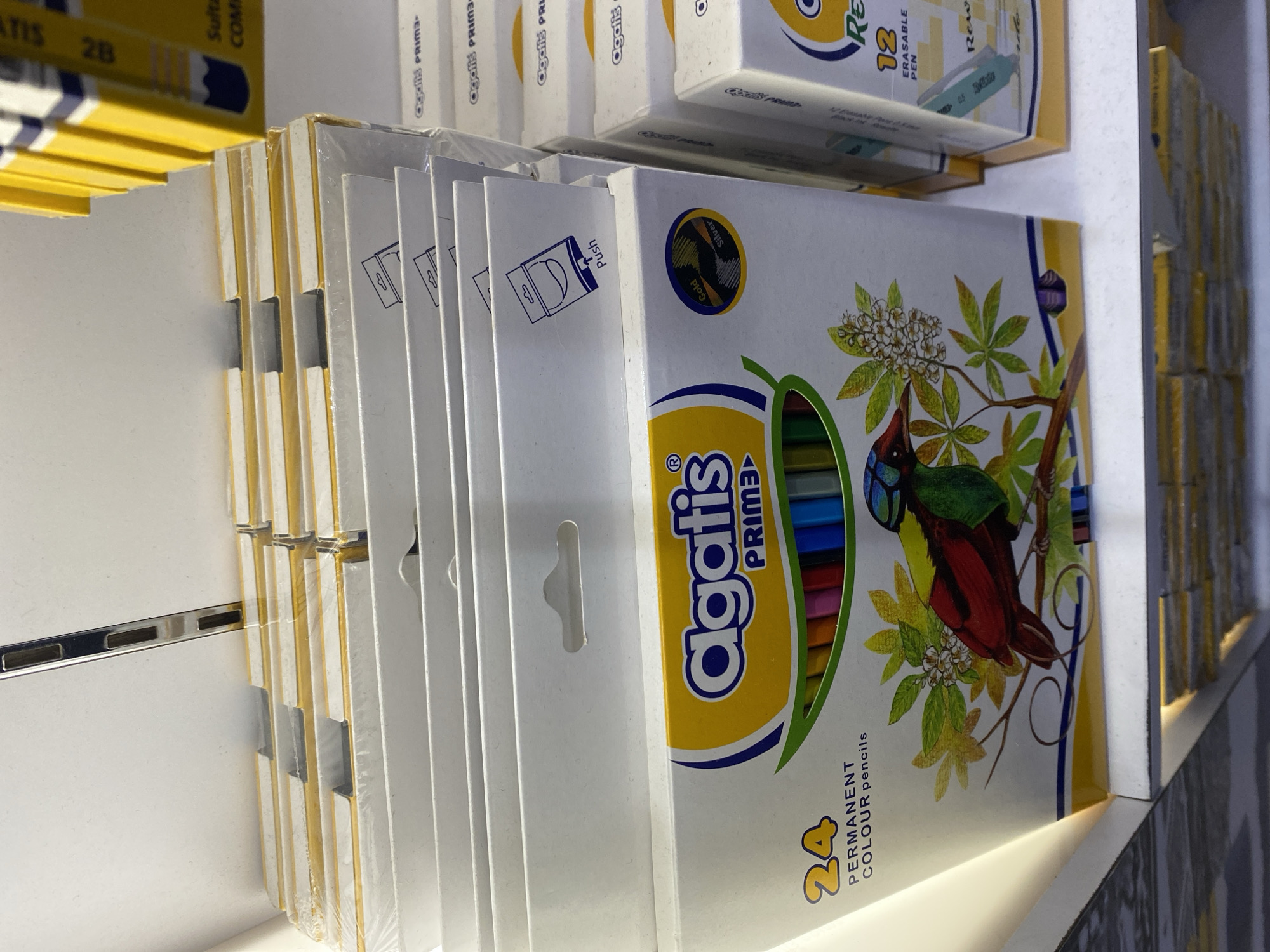 This image depicts a retail shelf in what appears to be a stationery store, possibly located in Depok, Indonesia, given the context. The shelf is stocked with several boxes of colored pencils and pens. The prominent item in the foreground is a box of "Agatis PRIME" permanent colored pencils, featuring an illustration of a bird and foliage. This box is partially wrapped in plastic and displays "24 PERMANENT COLOUR pencils" on its front.
Behind this box, other similar boxes are stacked, also appearing to be stationery products. To the right of the foreground box, another box labeled "Agatis PRIME" and containing "12 ERASABLE PEN" is visible. The shelves themselves are white with metal supports, characteristic of retail display units. The lighting suggests an indoor environment, likely a well-lit store. No people are present in the image, and the focus is entirely on the merchandise.