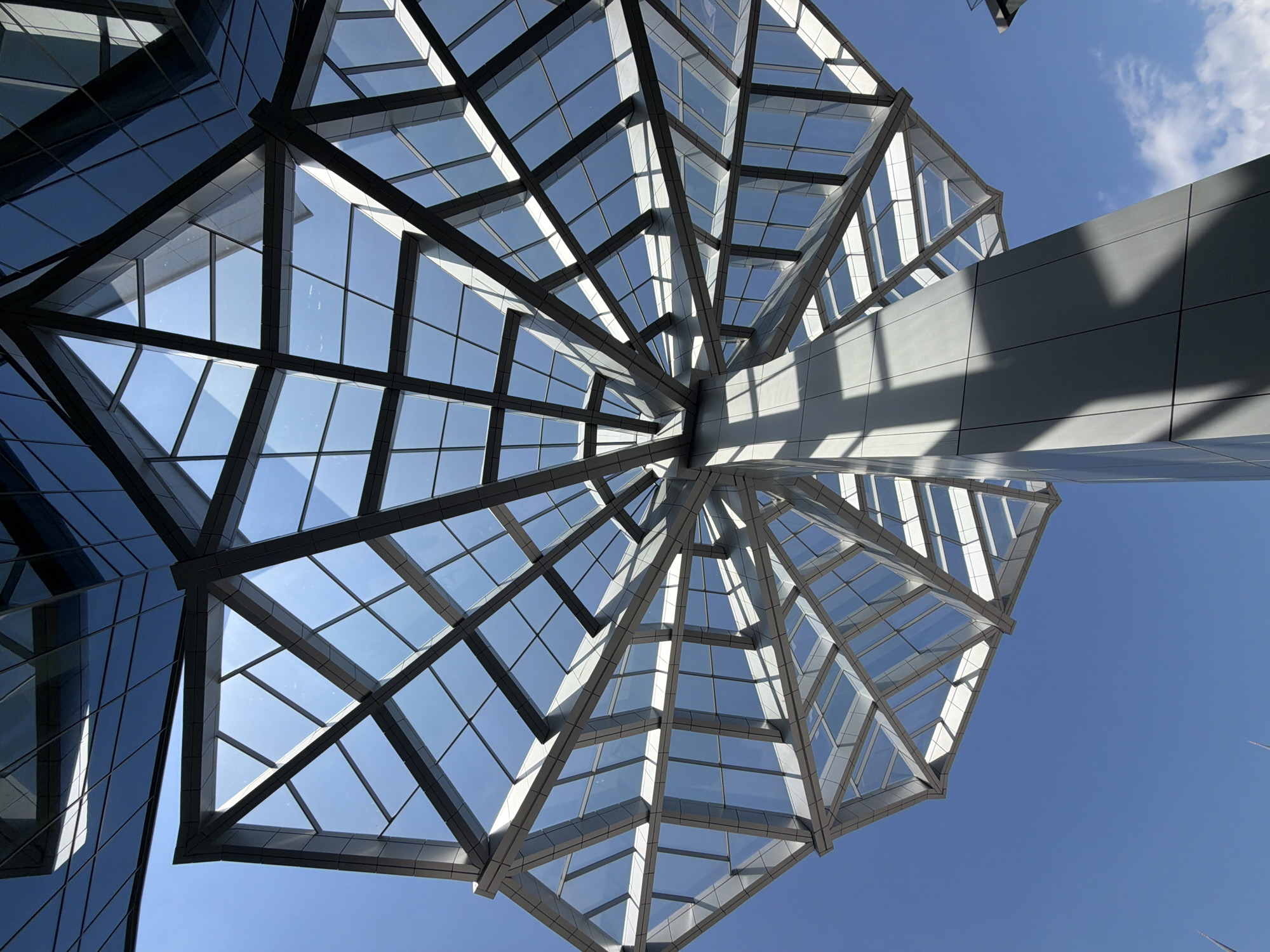The image shows a low-angle view of a modern architectural structure, likely a glass and metal canopy or roof element. The structure exhibits a radial pattern, with beams or supports emanating from a central point. The spaces between these supports are filled with panes of glass, allowing a view of the clear blue sky above. The canopy is attached to a building with a reflective glass facade on the left side of the image. A portion of a building or structure appears on the right, casting a shadow across the canopy. The location is unspecified and cannot be verified as Grozny, Russia from the visual information provided. No people, actions, or interactions are depicted.
