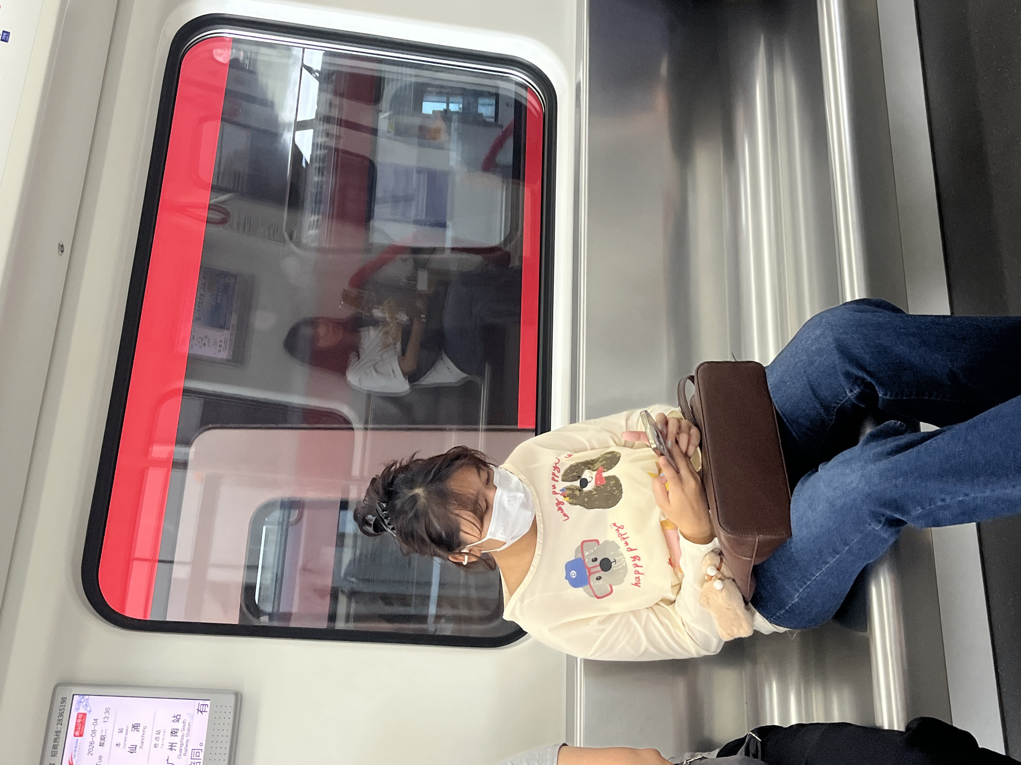 A young woman wearing a white face mask, a cream-colored long-sleeved shirt with dog graphics, and blue jeans sits on a metal bench inside a subway car. She is looking down at her phone, which she holds in her hands. A brown purse rests on her lap, and a small, fluffy keychain is attached to it. The window to her right reflects another passenger in the car. A digital display board on the left side of the image shows the date as 2026-08-04 and indicates the current station as Xianzhong in Shunde District, China, with Guangzhou South Railway Station as the terminal. The subway car interior is clean and modern, with silver-colored seats and a red trim along the window.
