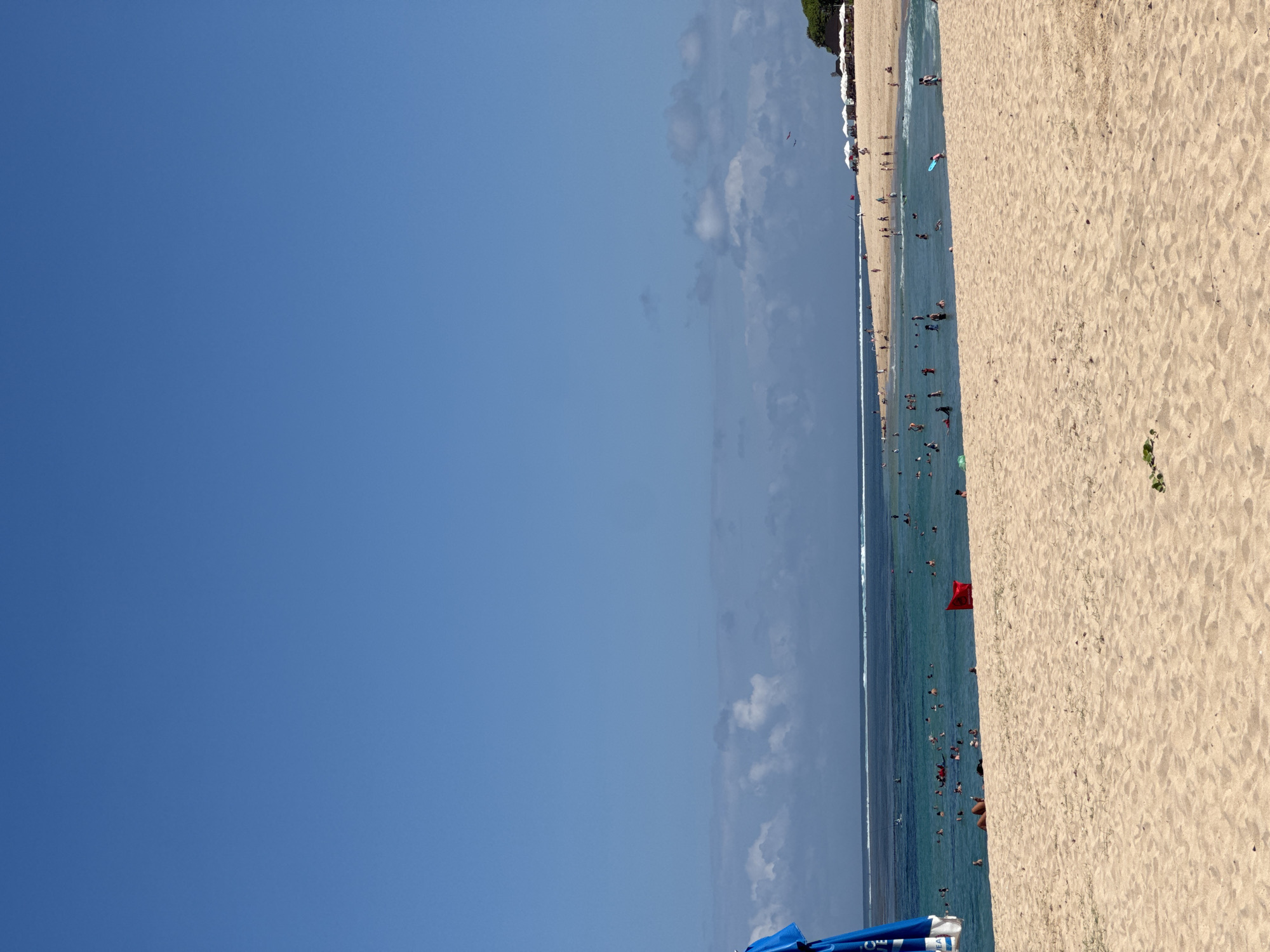 The image captures a wide expanse of a sandy beach on a clear, sunny day in Kuta Selatan, Indonesia. The foreground is dominated by light-colored sand with sparse patches of dry grass. In the middle ground, the turquoise ocean is filled with numerous people swimming and wading. A red flag is planted in the sand near the water's edge, possibly indicating a warning or a designated swimming area. Further out, the ocean meets the horizon under a bright blue sky, with a thin line of clouds visible. On the right side of the middle ground, a line of white umbrellas suggests a resort or beachside establishment. In the far distance, a lush green tree line borders the beach. A blue umbrella is partially visible in the left foreground, obscuring the very edge of the frame.