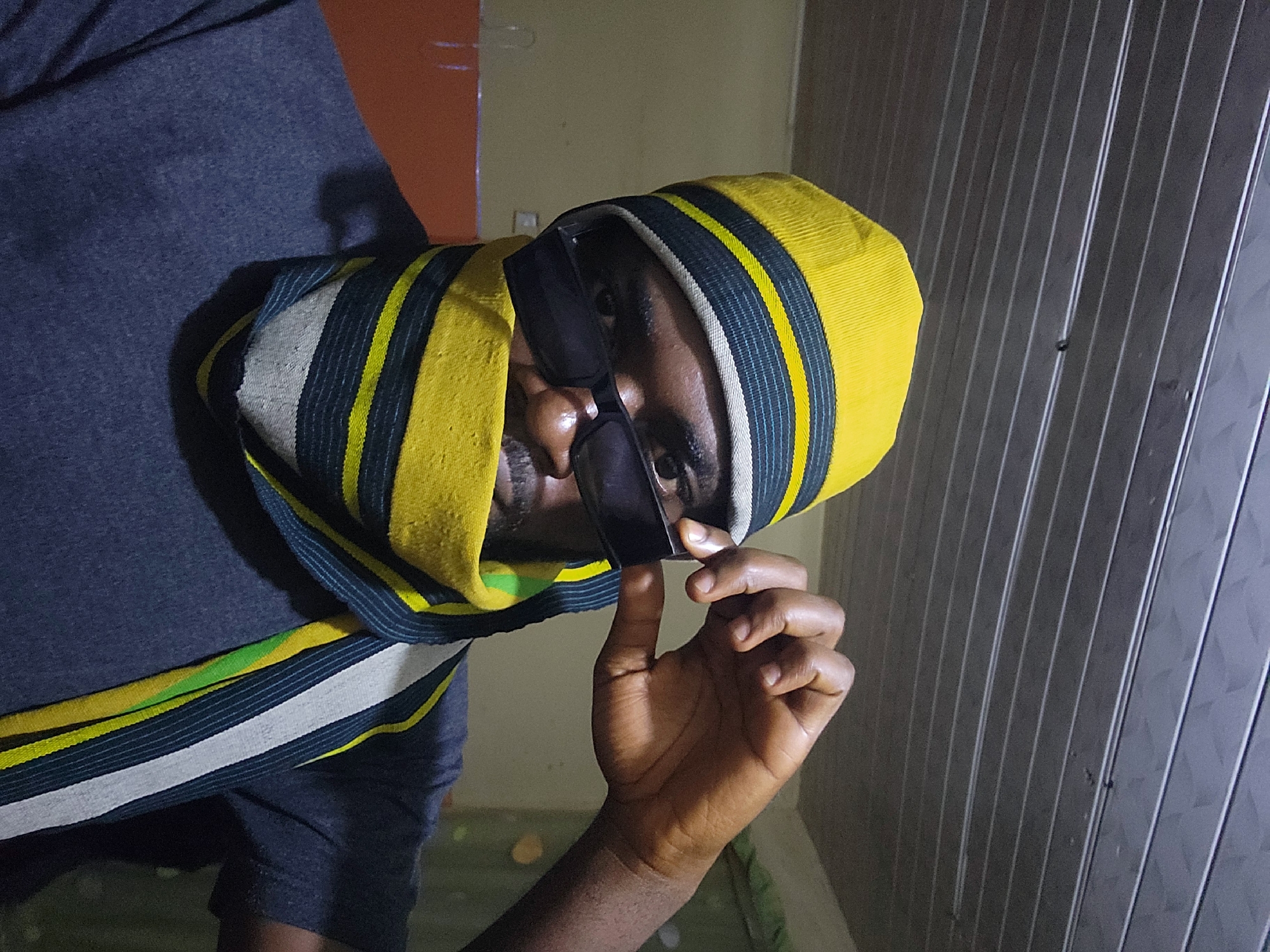 A medium shot shows a person's head and shoulders from a low angle. The person is wearing a yellow and striped beanie, sunglasses, and a scarf. The scarf is wrapped around the person's neck and lower face, with its ends hanging down over their chest. The person's right hand is raised, and their fingers are touching the side of their sunglasses. The background appears to be an indoor setting with a textured ceiling and a plain wall. The lighting is dim, casting shadows on the person's face. There are no clear indicators of the specific location in Jos, Nigeria, beyond the implied presence of the person.