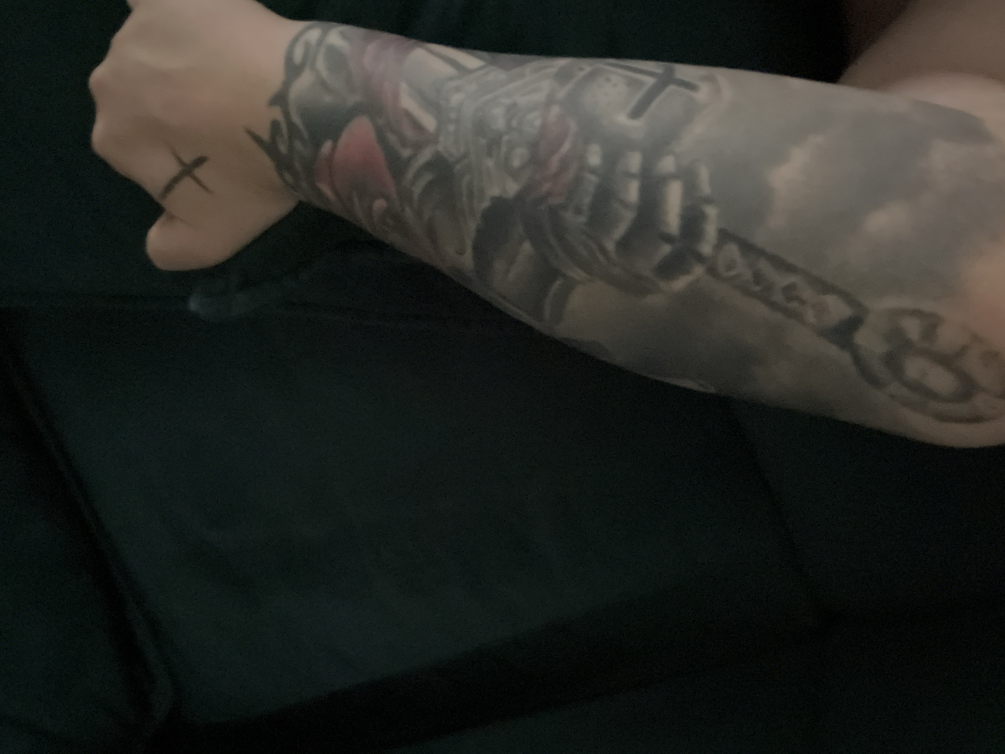 The image shows a person's arm covered in tattoos, resting on a dark green upholstered surface. A black 'X' tattoo is visible on the back of the hand, near the index finger. The tattoos on the arm appear to depict a stylized skull with red accents and a long, ornate object resembling a sword or scepter, set against a background of clouds. The lighting is dim, casting shadows on the arm and the surrounding furniture. The image does not contain any elements that would allow for the identification of a specific location in the city of Varna, Bulgaria.