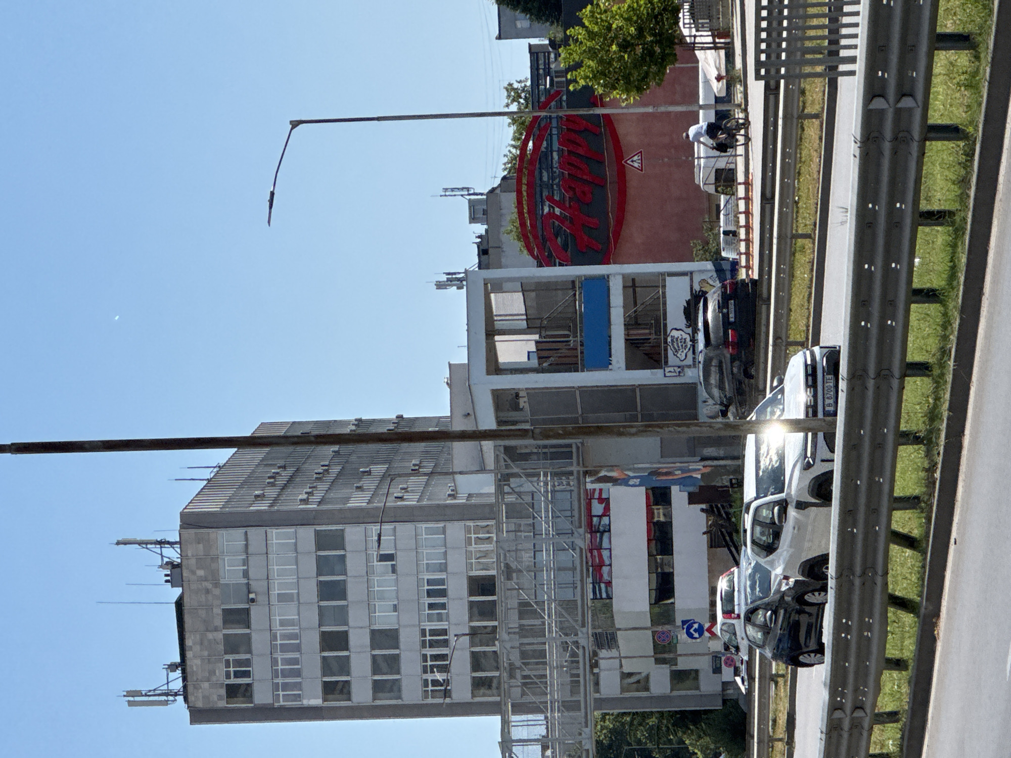 The image captures a view of a street scene in Varna, Bulgaria, on a bright, sunny day. In the background, a tall, grey, multi-story building with many windows dominates the skyline. Several antennas are visible on its roof. Adjacent to this building is another structure with a large, red, circular sign featuring the word "Happy" in bold, red lettering. A street light on a tall pole stands prominently in the foreground, partially obscuring the view of the buildings.
In the mid-ground, a pedestrian bridge crosses over the street. Below this bridge, on the side of the "Happy" building, there appears to be some graffiti or artwork. Cars are visible on the road, including a dark-colored car and a white SUV with its license plate partially visible. A person on a bicycle is seen on the right side of the image, riding along the road. A traffic barrier with a reflective surface separates the lanes of traffic. The environment appears to be urban, with green grass and trees visible on the sides of the road. The sky is clear and blue.