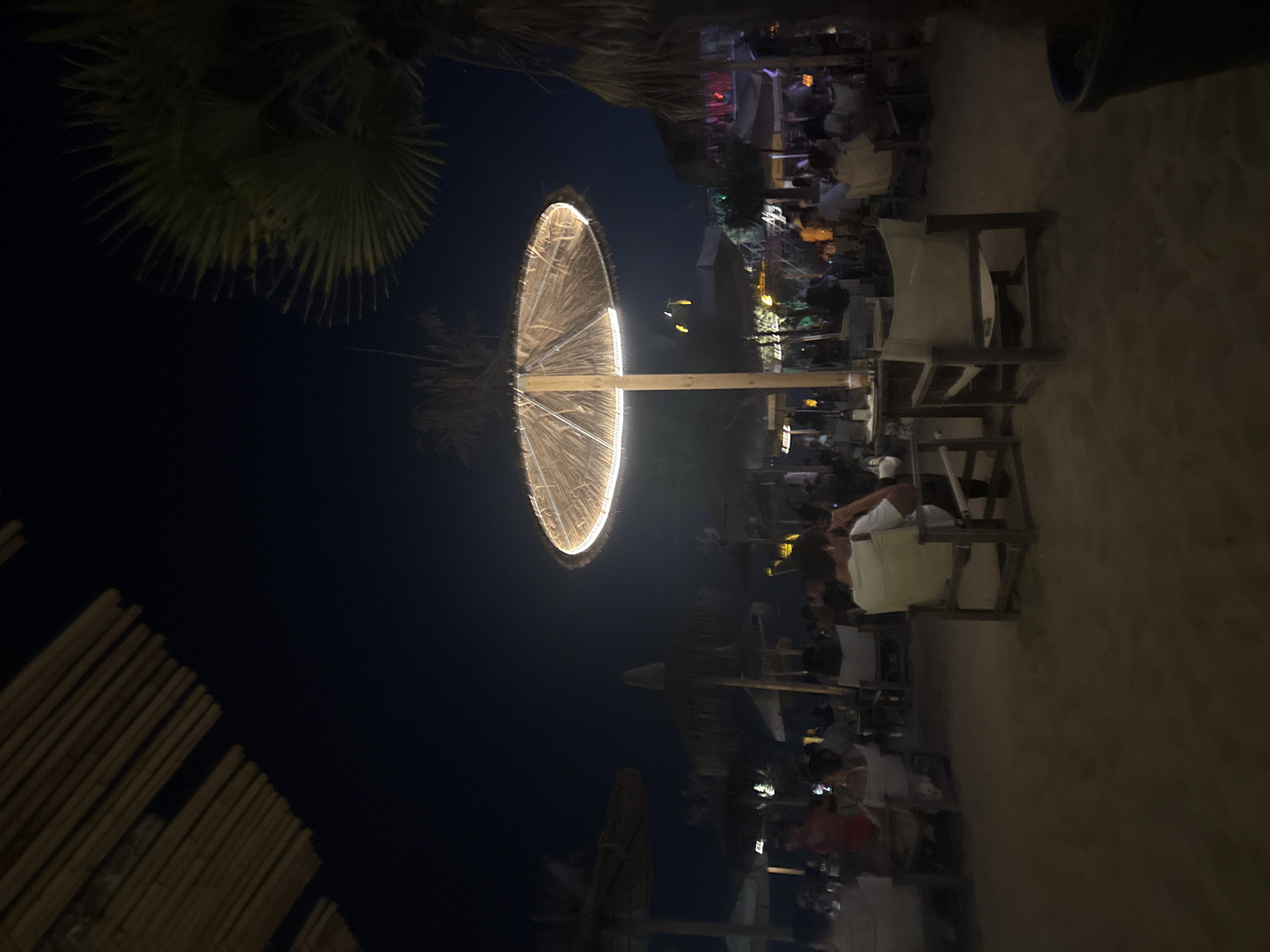 The image depicts a nighttime scene on a sandy beach, likely a resort or outdoor dining area. Several straw-covered umbrellas are spread across the sand, some illuminated by string lights. In the foreground, two chairs are positioned at a table, one occupied by a person from behind, wearing a light-colored t-shirt. Other tables and chairs are visible, with groups of people seated and dining. Palm trees are present, adding to the tropical ambiance. The sky is dark, indicating it is night. The lighting is dim, with artificial lights from the umbrellas and surrounding establishments illuminating the scene. The overall impression is of a relaxed and social evening at a beachside venue.