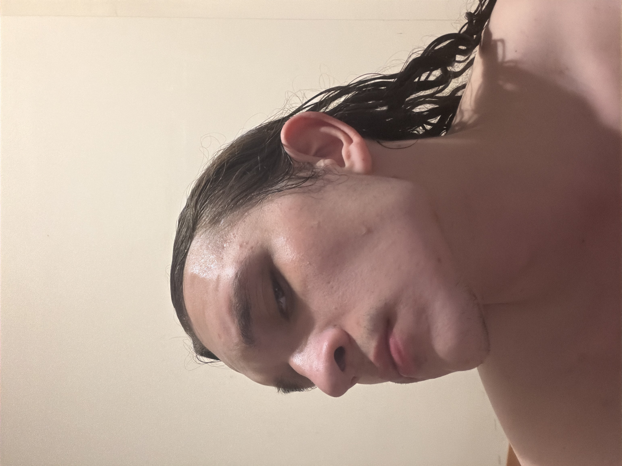 A close-up, rotated portrait features the head, neck, and upper shoulder of a young individual, likely male, against a plain, light-colored wall. The subject's head is tilted sharply to the left, with their face angled downwards and slightly away from the camera. Their dark, wavy hair appears wet, clinging to the scalp and neck. The skin, particularly on the forehead and nose, shows a glossy sheen, suggesting dampness or perspiration. The visible ear is distinct. The eyes are partially closed or cast downwards, giving a neutral or contemplative expression. The background is a uniform, light cream or beige wall, indicating an indoor setting. The lighting is soft and even, typical of indoor artificial light. The image captures the subject in what appears to be a post-shower or post-washing moment. No specific objects or environmental details are visible to further define the setting. The given location context is Plovdiv, Bulgaria, though no visual cues within the image confirm this. There is no visible text.